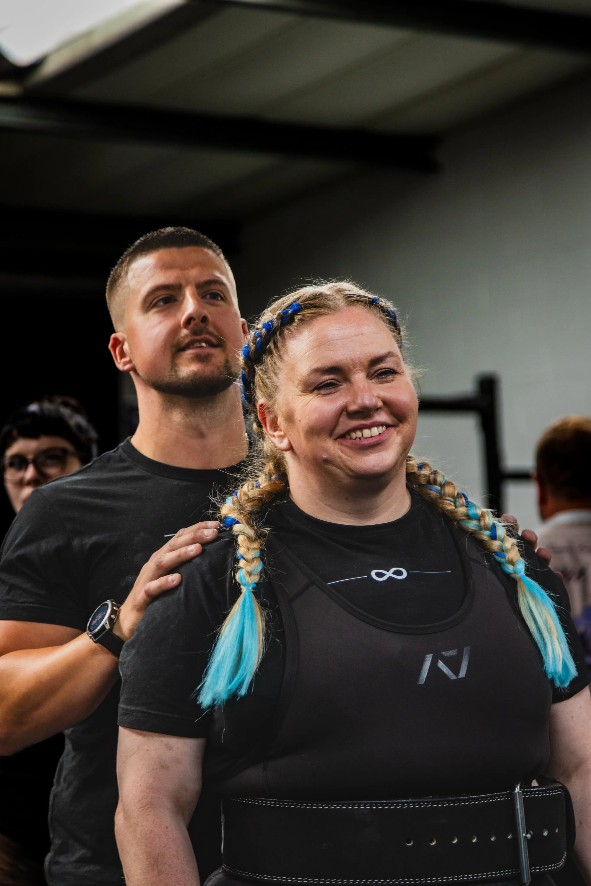 A group of people, including a woman with blue and blonde braided hair, standing closely together indoors, with one person resting a hand on the woman's shoulder, likely during a group activity or event.