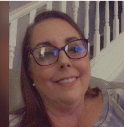 A woman with glasses smiling, sitting on a couch in front of a white staircase railing.