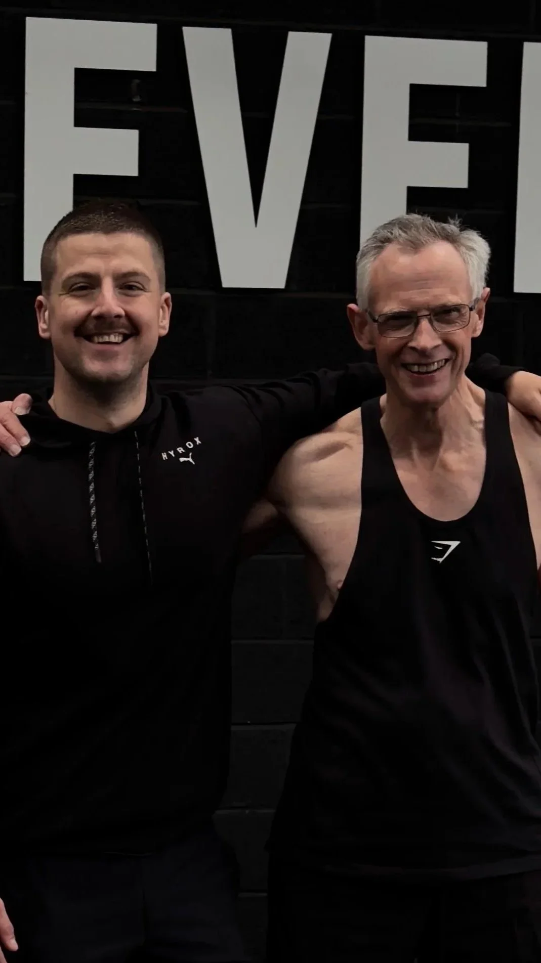 Two smiling men standing next to each other with arms around each other, in a gym. The younger man on the left is wearing a black hoodie, and the older man on the right is wearing a black tank top, glasses, and has gray hair. There is a large sign with the partially visible word 'FIVE' behind them.