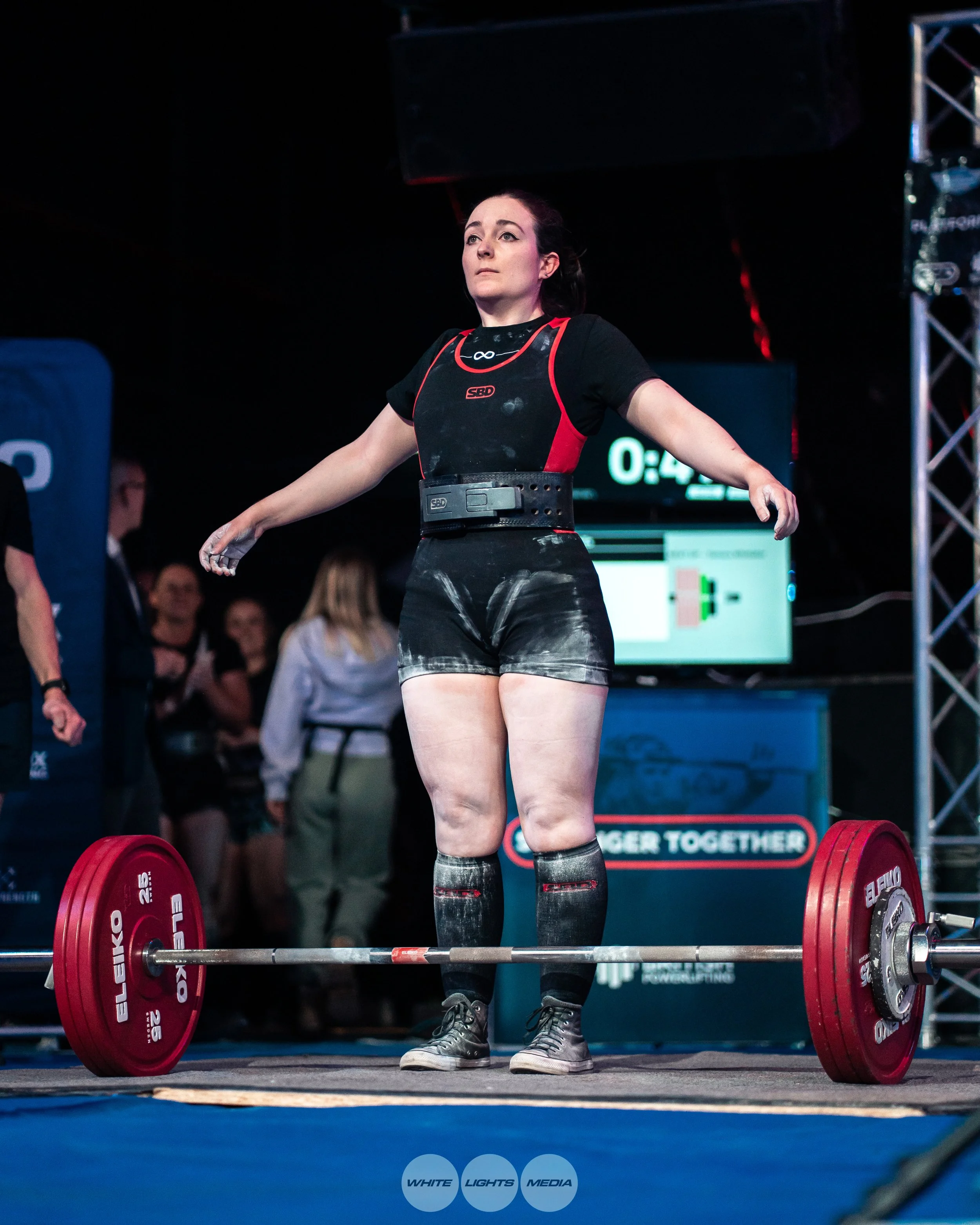 A female powerlifter standing on a weightlifting platform with arms outstretched, wearing a black and red competition singlet, a weightlifting belt, and knee-high socks. Red weight plates are on a barbell on the ground in front of her, and an electronic scoreboard and audience are visible in the background at a weightlifting competition.