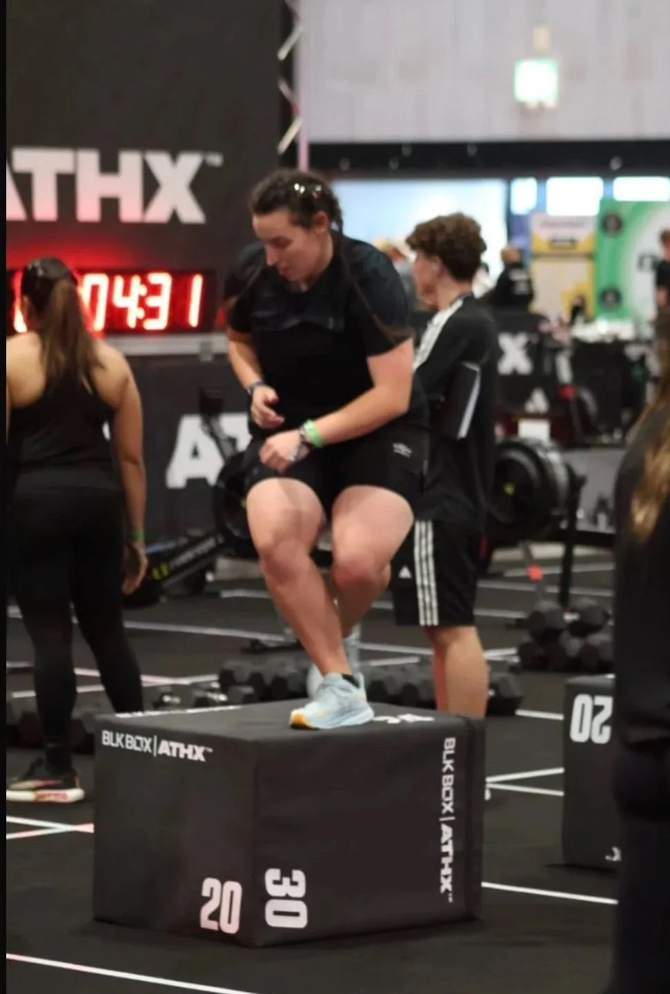 A woman preparing to do a box jump at a fitness event. She is standing on a black padded box with white and pink markings, in a gym environment with other participants and equipment visible.