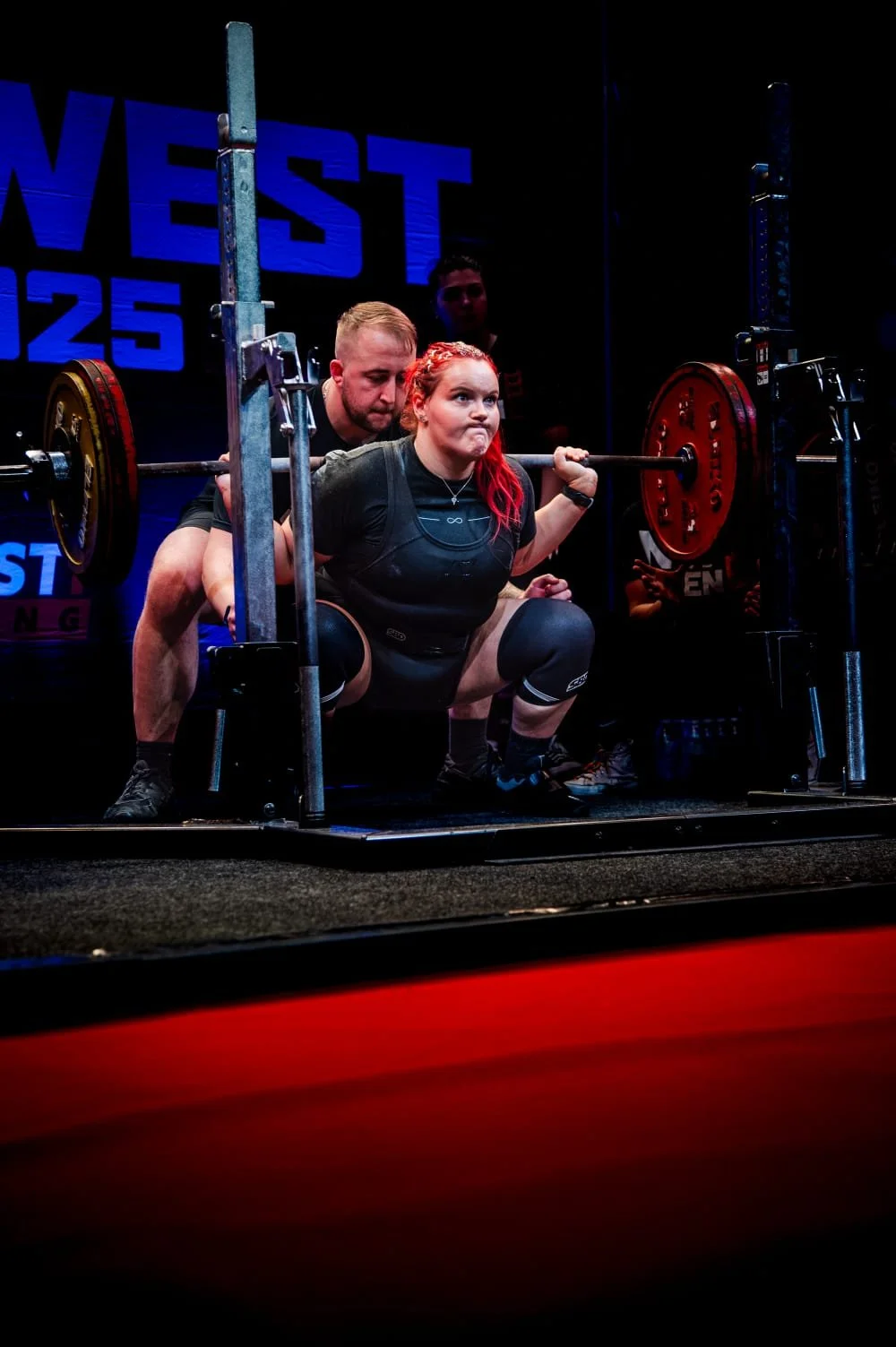 Woman doing a squat with a barbell during a powerlifting competition, with spotters helping her.