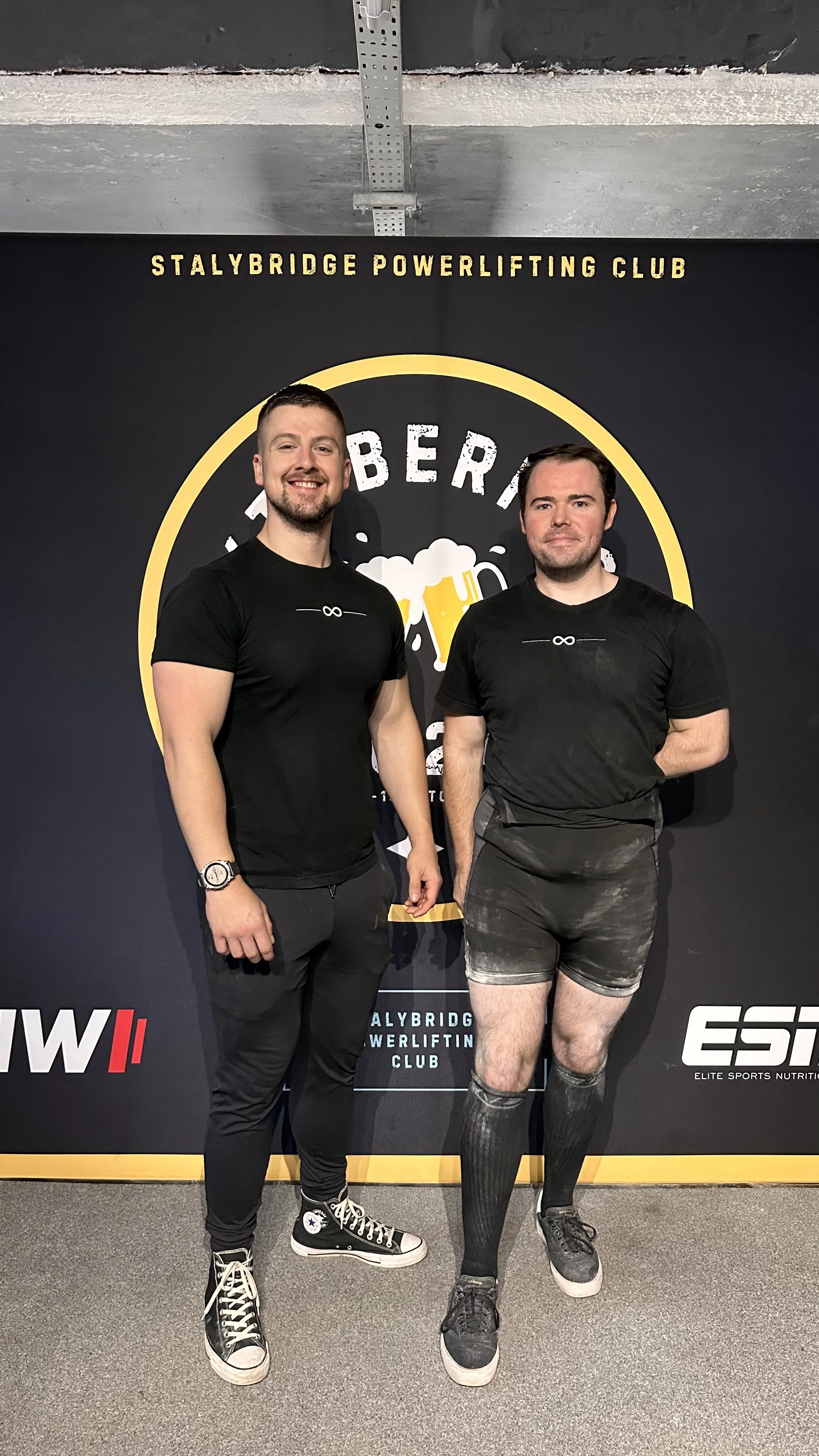 Two men standing in front of a black backdrop with the words "Stalybridge Powerlifting Club" and a logo of a beer mug. The man on the left is smiling, wearing a black T-shirt, black pants, and black Nike high-top sneakers. The man on the right is standing with a neutral expression, wearing a black T-shirt, gray shorts, and black athletic socks and shoes.