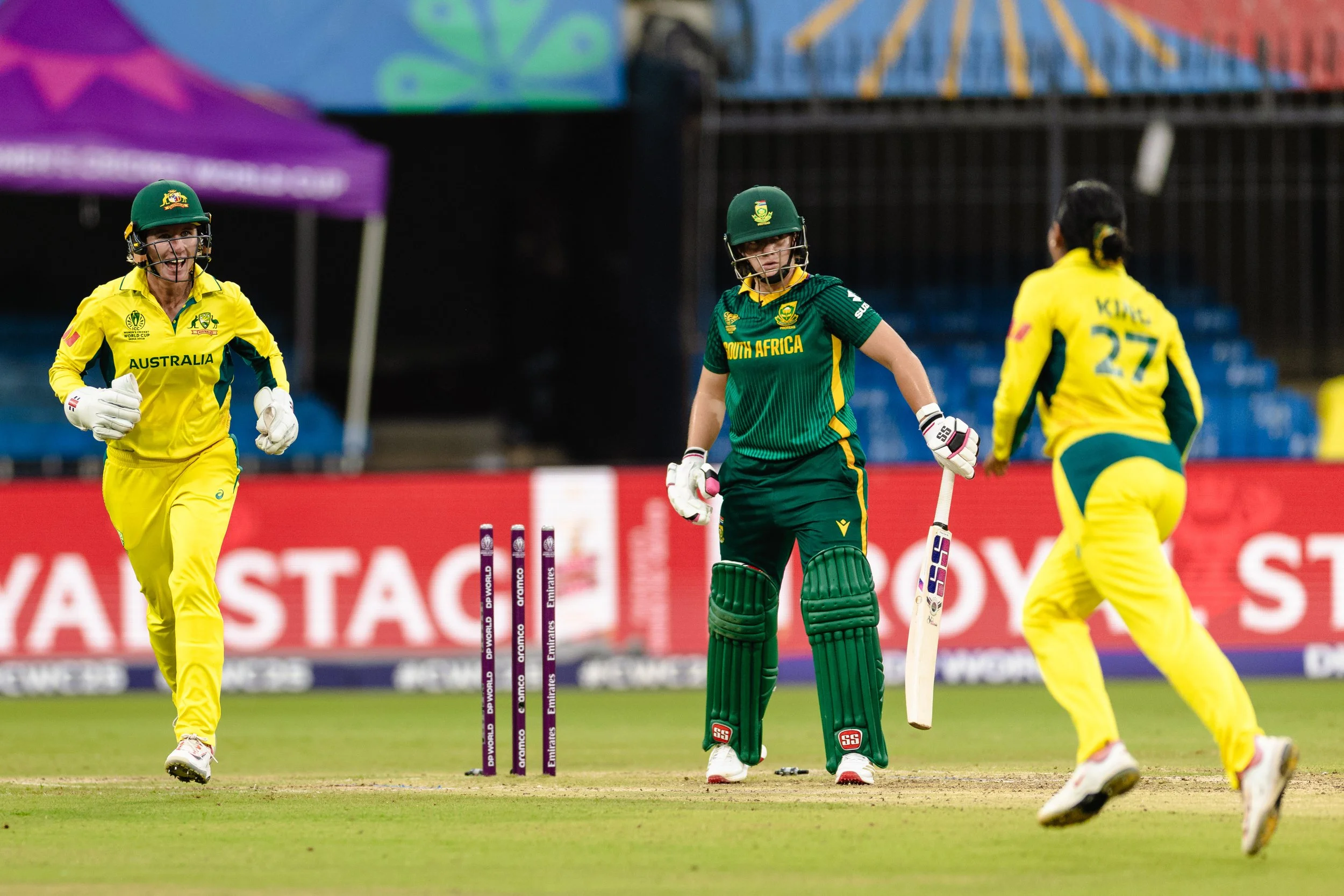 Cricket players from Australia and South Africa on the field during a match, with some players wearing yellow and some in green uniforms, and cricket stumps in the background.