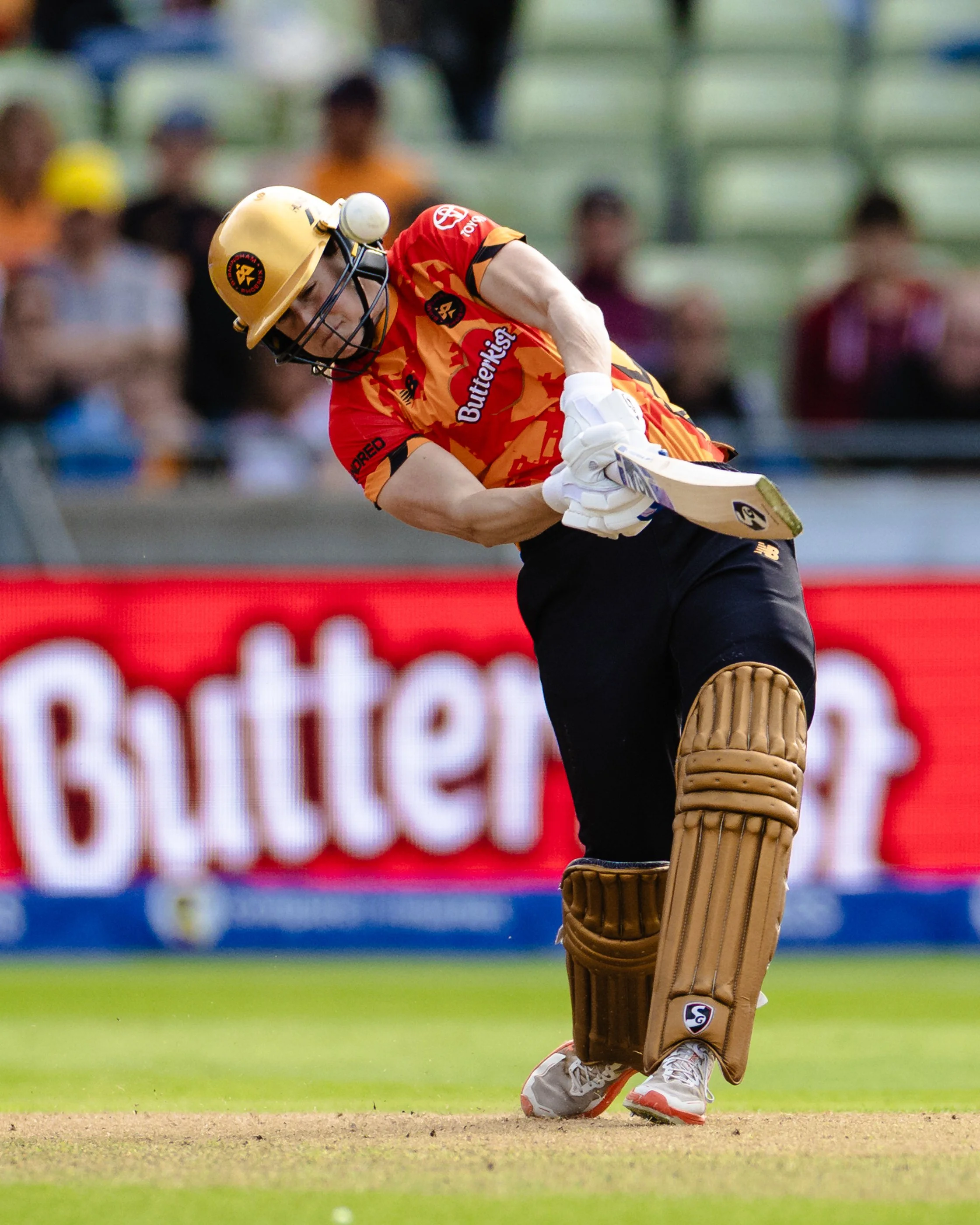 Cricket player wearing a yellow helmet and red and orange uniform, holding a cricket bat, on the field with spectators blurred in the background.