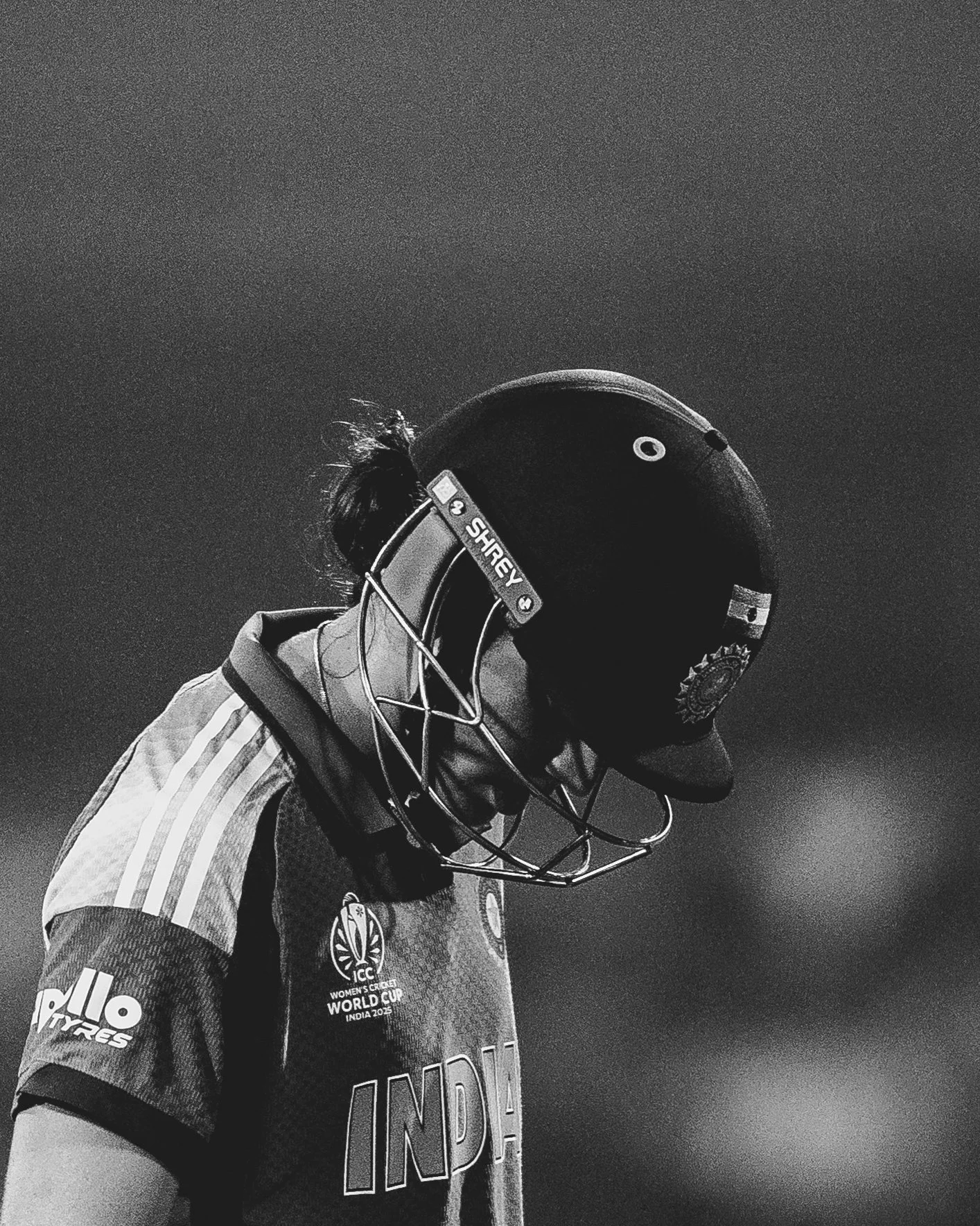 A black and white photo of a female cricket player looking down, wearing a helmet, cricket uniform with logos, and what appears to be a skull emblem on the shirt. The helmet has a face guard and the shirt mentions ICC Women's Cricket World Cup India 