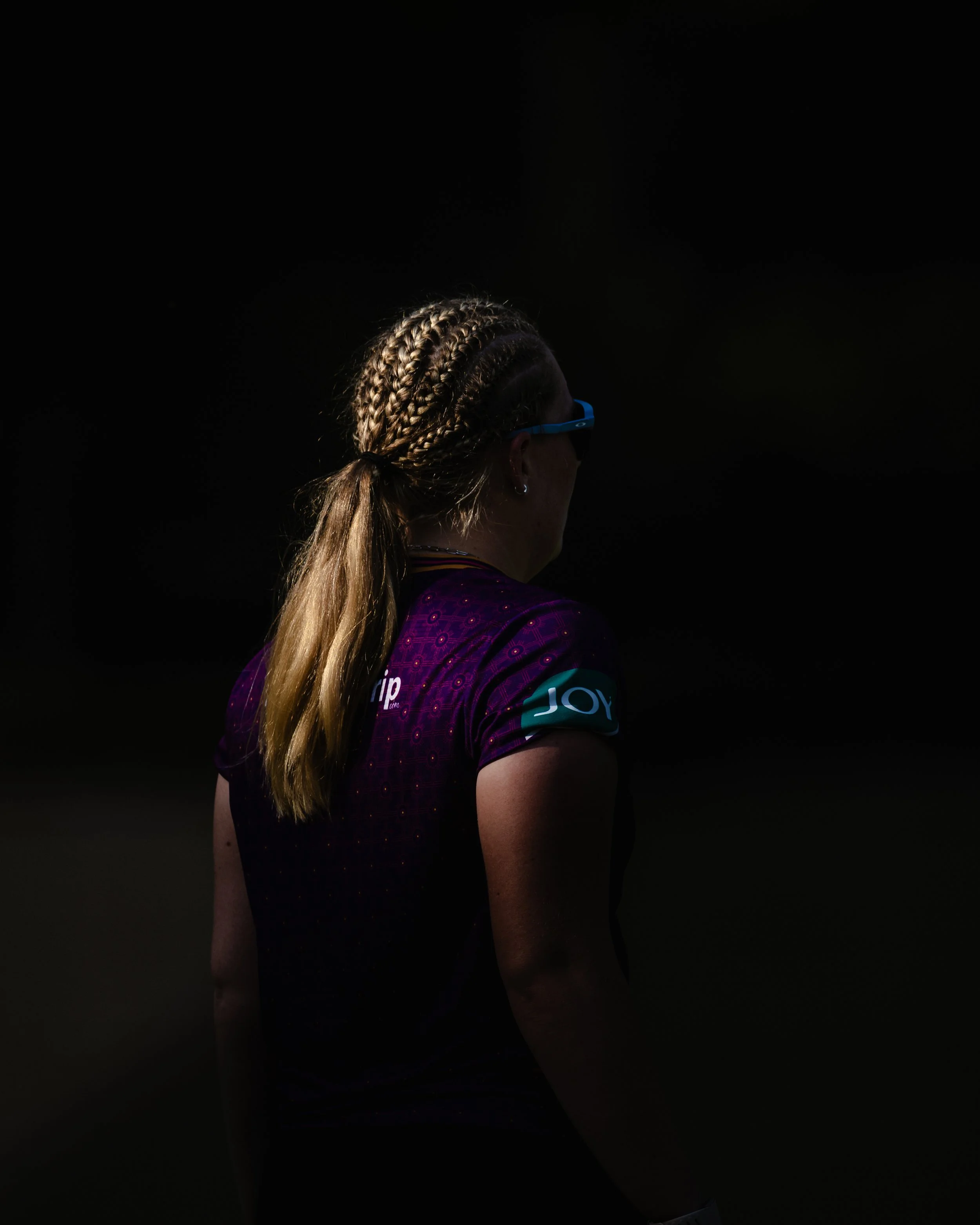Side view of a woman with blonde braided hair wearing blue glasses and a purple sports jersey with sponsor logos, standing outdoors at night.