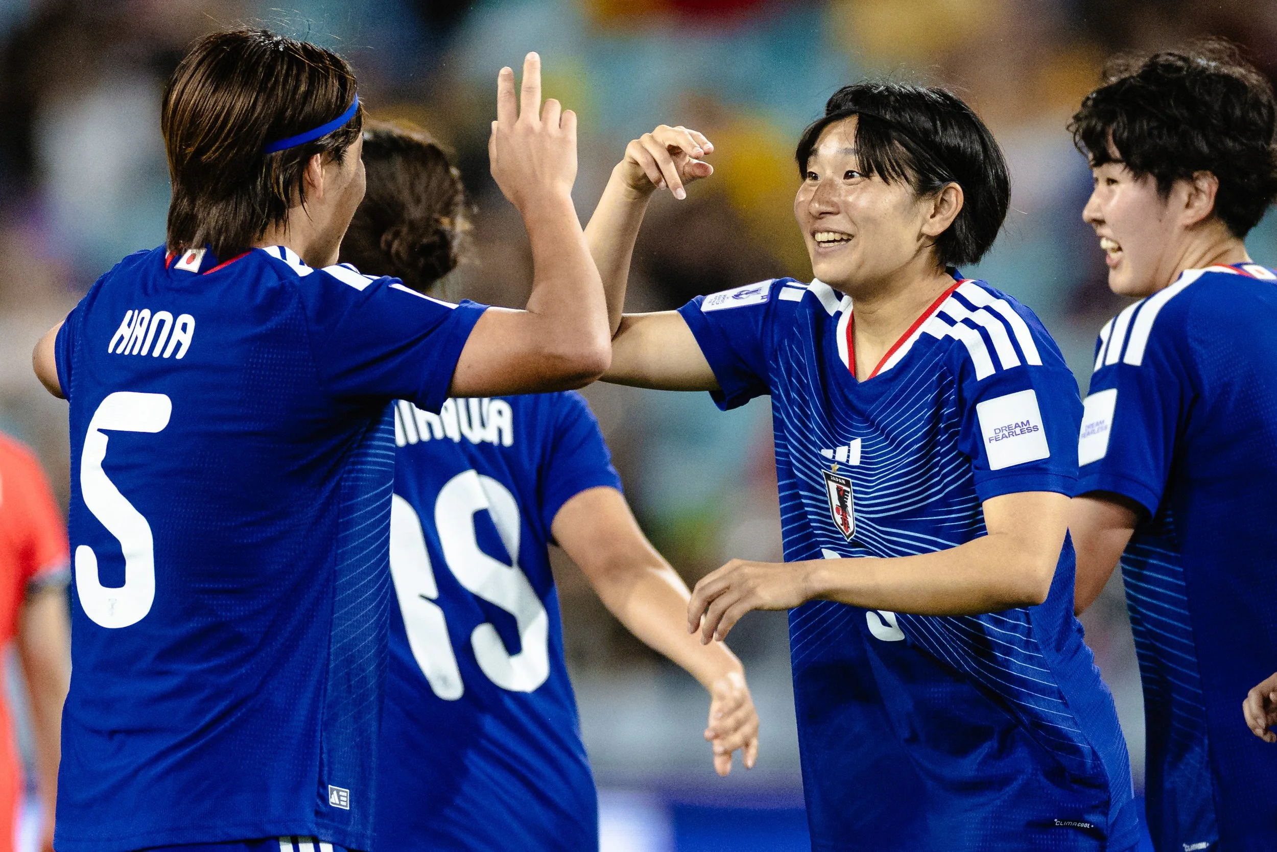 Four female soccer players in blue jerseys are celebrating on the field, smiling and greeting each other with a high five.