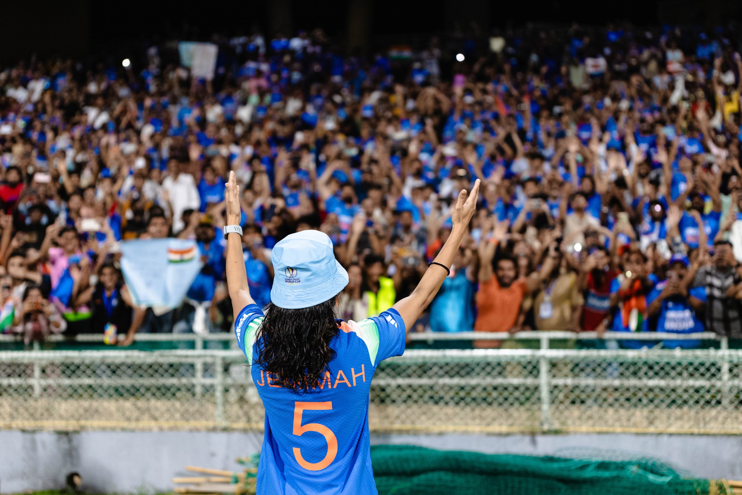 A woman wearing a blue cricket jersey with the number 5 and the name 'JEMMAH' on the back, raising her arms in front of a large crowd at a cricket stadium, with many fans wearing blue and holding signs.