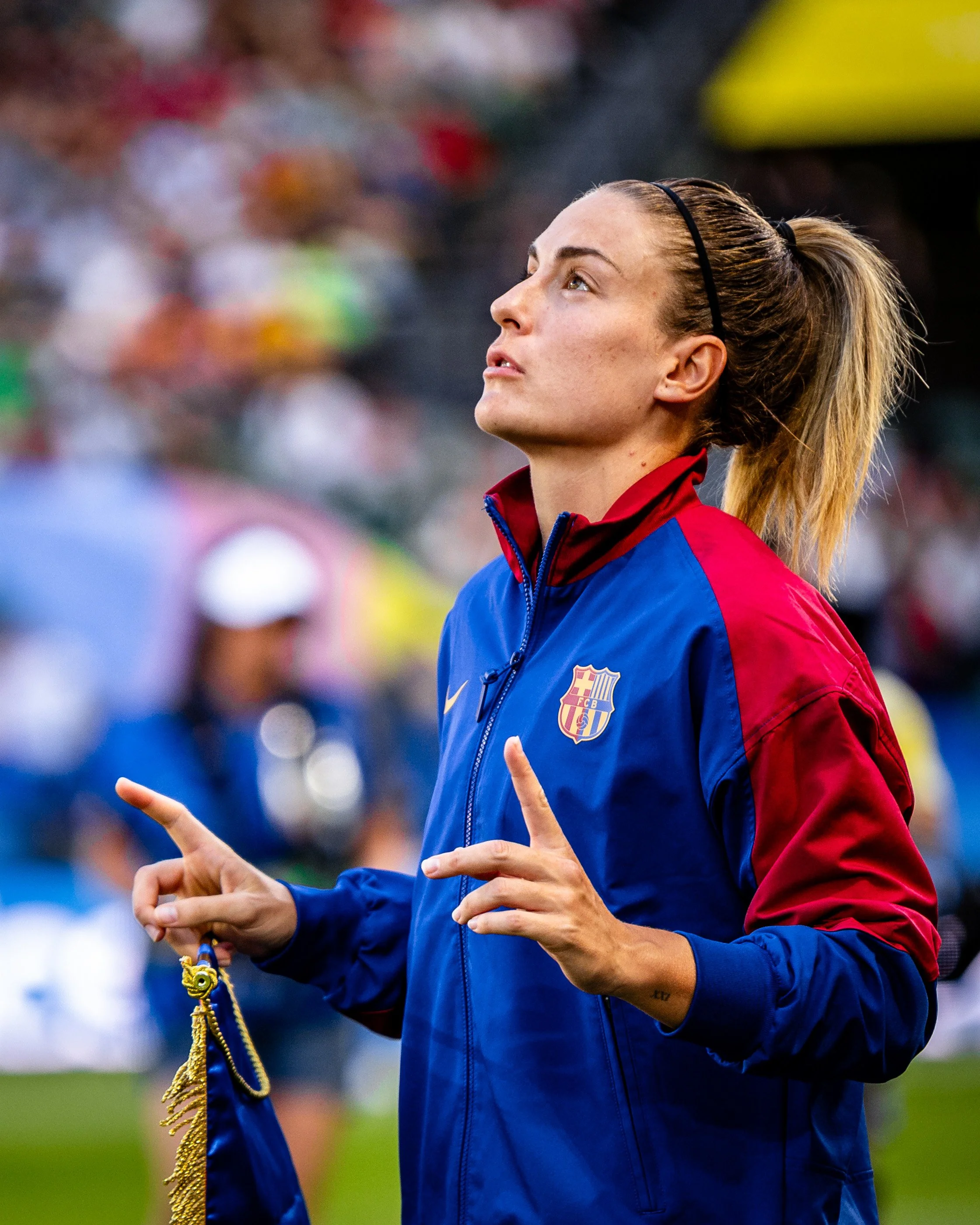 A female athlete from FC Barcelona on a sports field, holding a medal and ribbon, wearing a blue and red jacket with the team badge visible, looking upward with a focused expression.