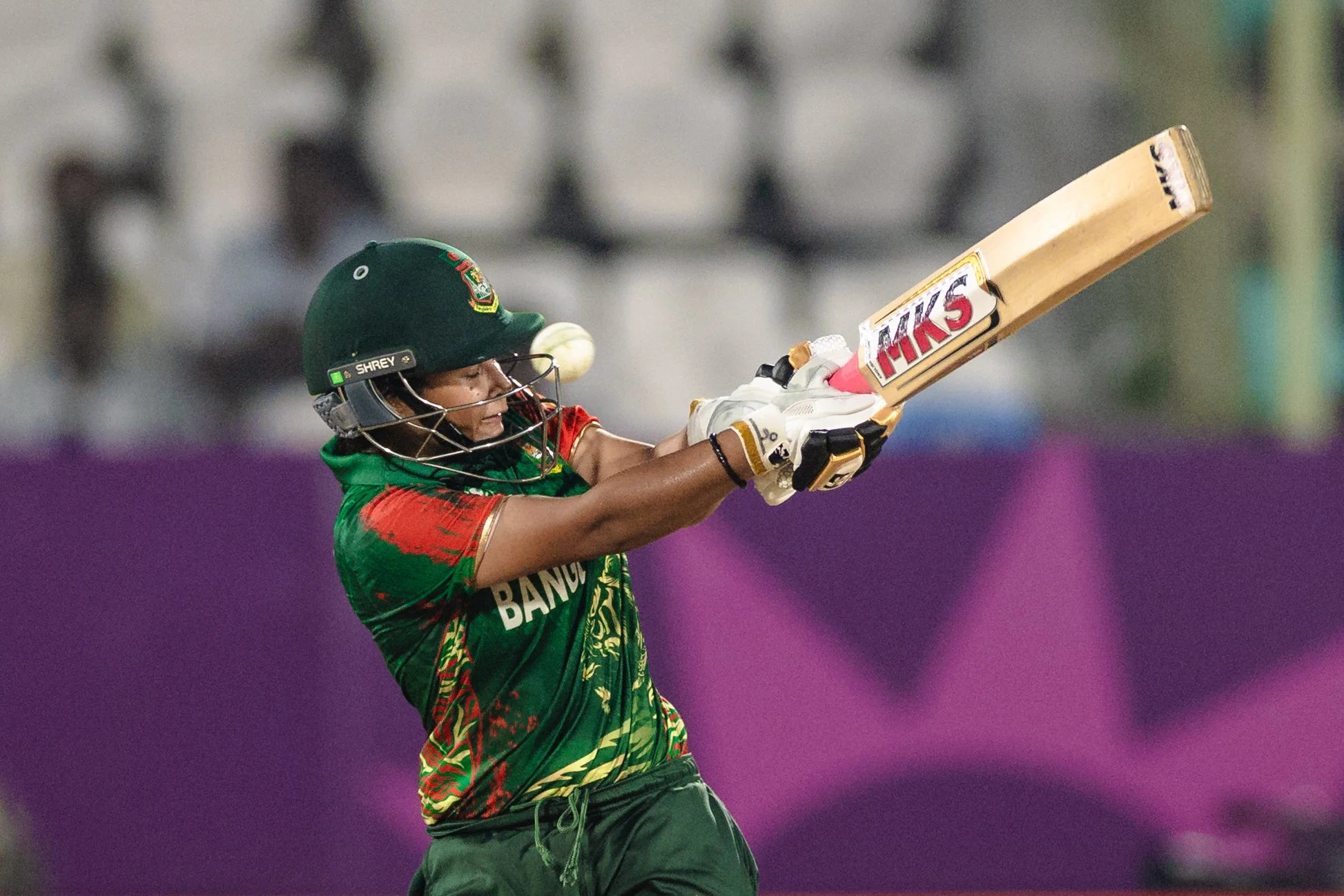 A cricket player from Bangladesh hitting a ball during a match, wearing a green helmet and jersey with red and green patterns.
