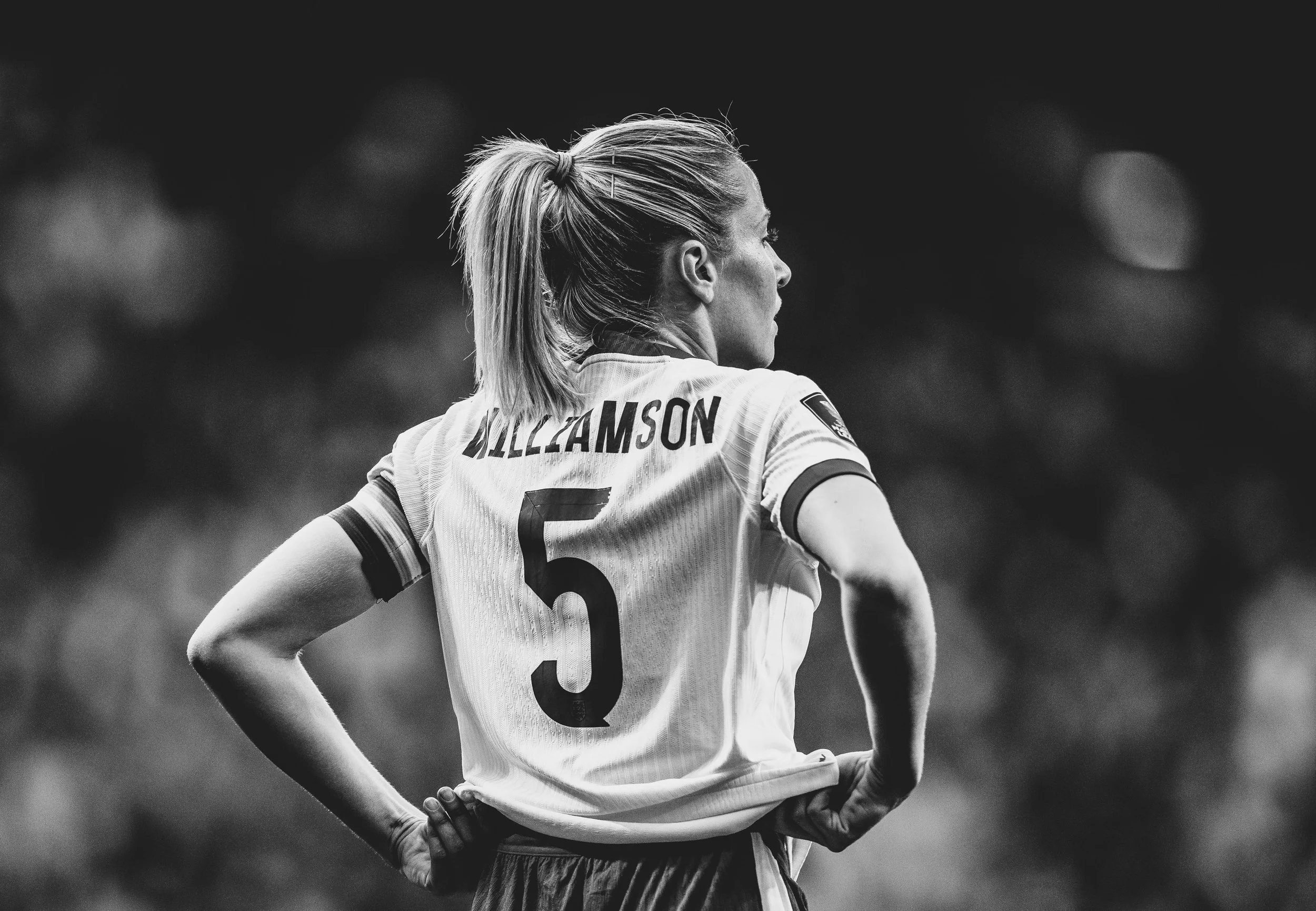 A black and white image of a female athlete from behind, with her hair in a ponytail, wearing a sports jersey with the name 'WILLIAMSON' and the number 5 on the back, standing with her hands on her hips during a sports game.