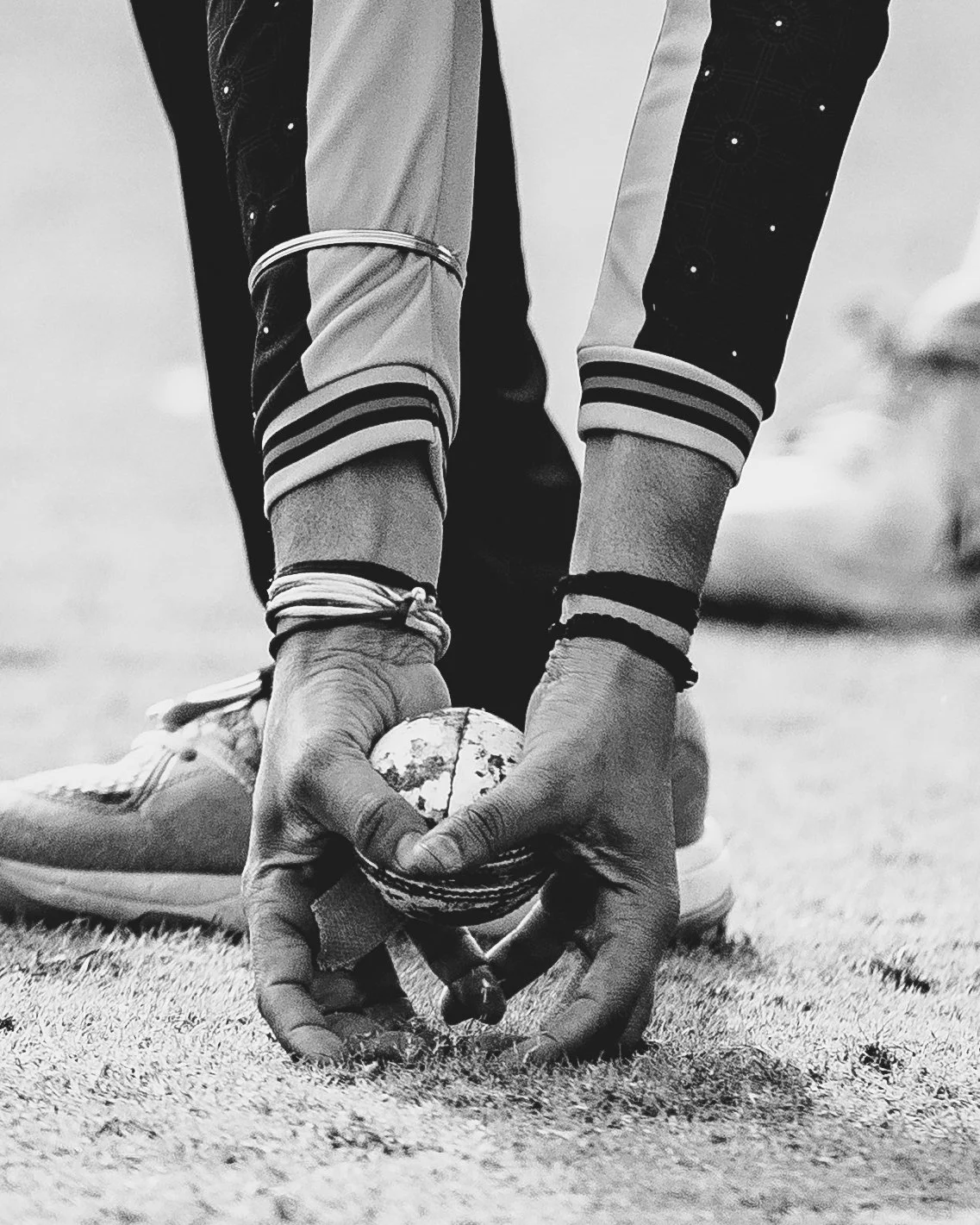 A person kneeling on a sports field holds a worn tennis ball in their hands, with one foot slightly in front of the other and wearing athletic shoes and striped wristbands.