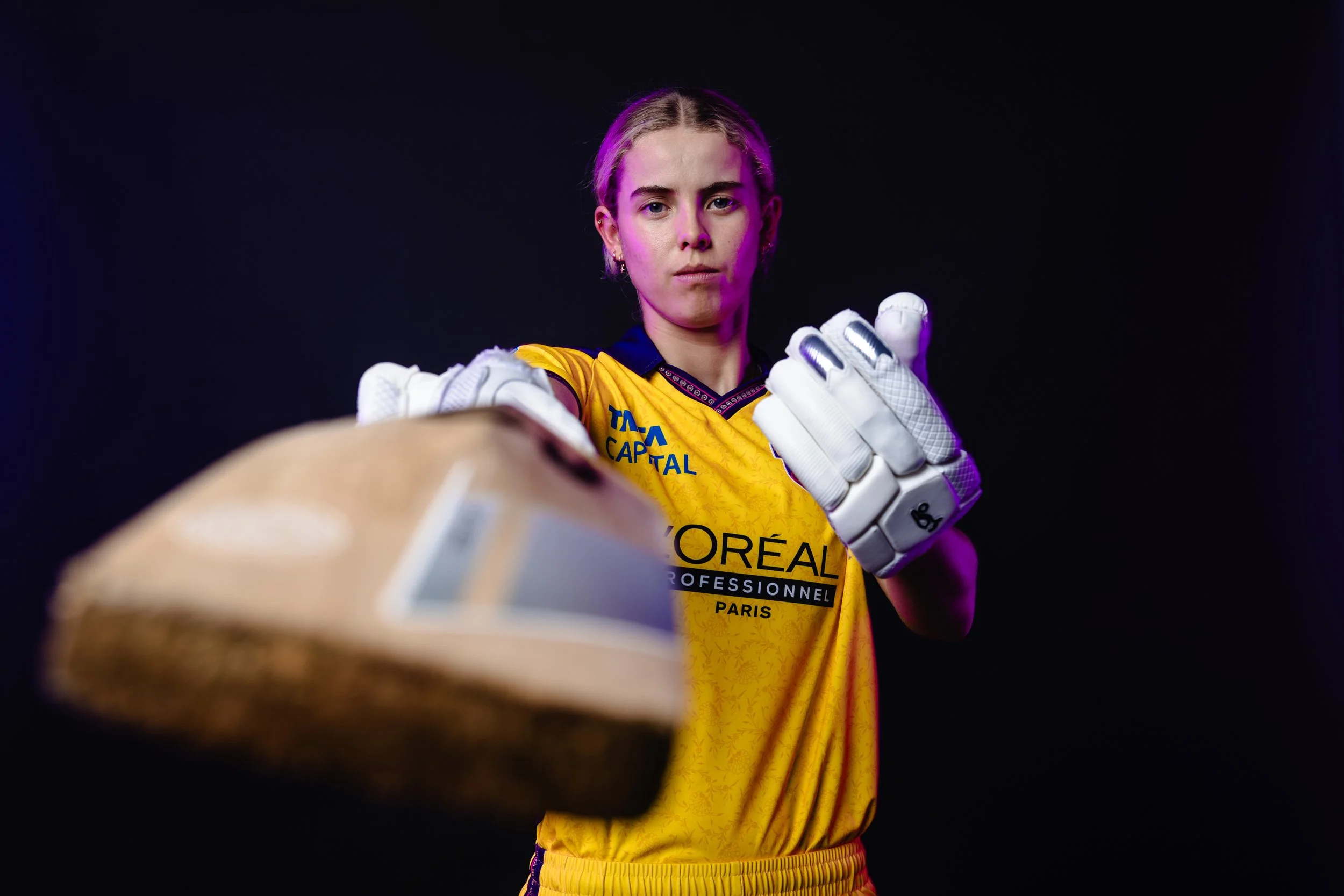 A young woman in a yellow sports jersey and white goalkeeper gloves reaching out with her hand towards the camera, with a serious expression on her face, against a dark background with purple lighting.