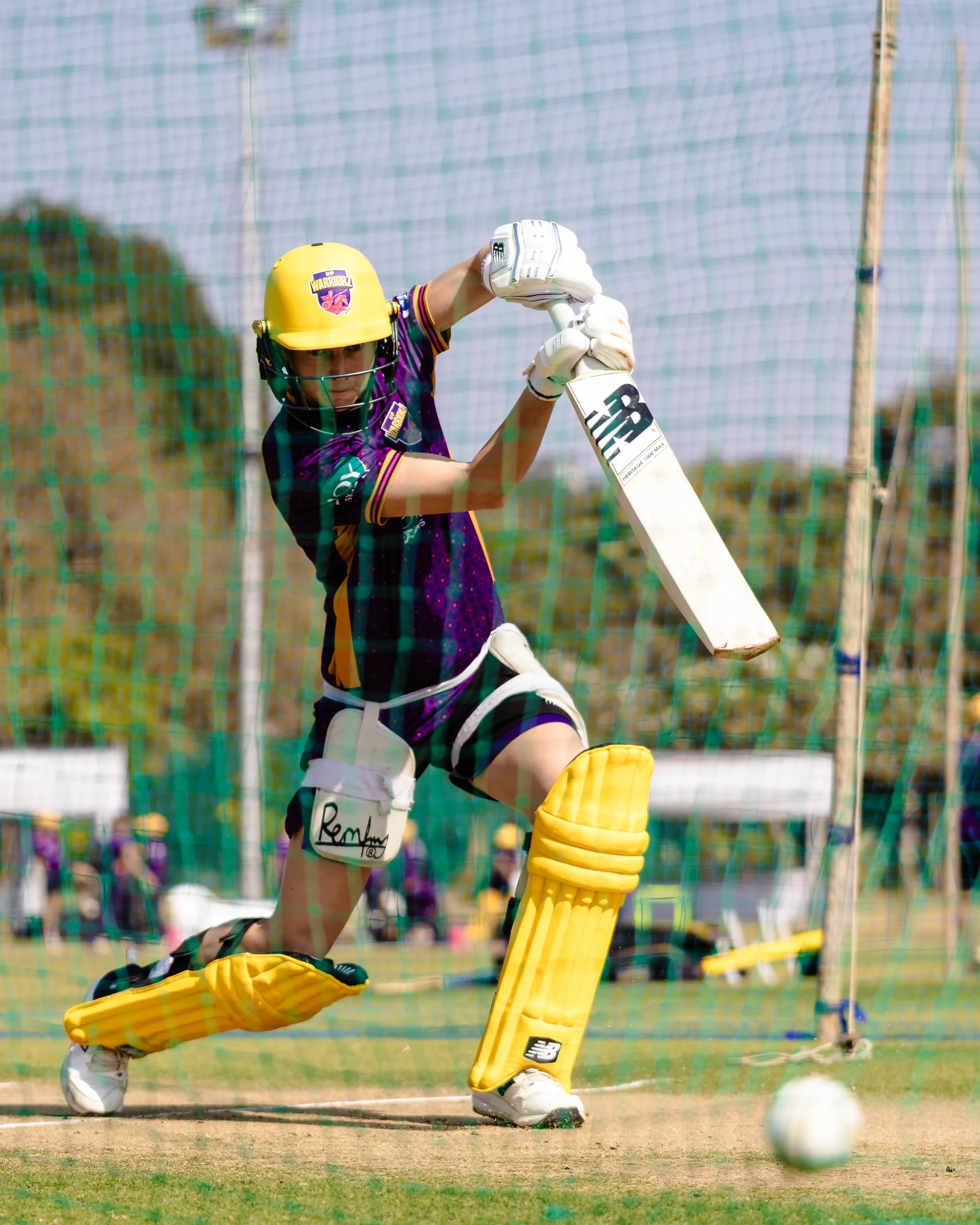A cricket wicketkeeper is fielding in the cricket match, wearing a yellow helmet with a logo, white gloves, yellow leg pads, and holding a cricket bat with a logo, behind a yellow wicket in the cricket field.
