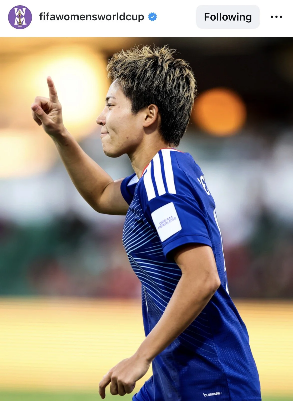A female soccer player in a blue jersey with white stripes and the number 16 on her back, smiling and pointing upwards with her right hand during a game.