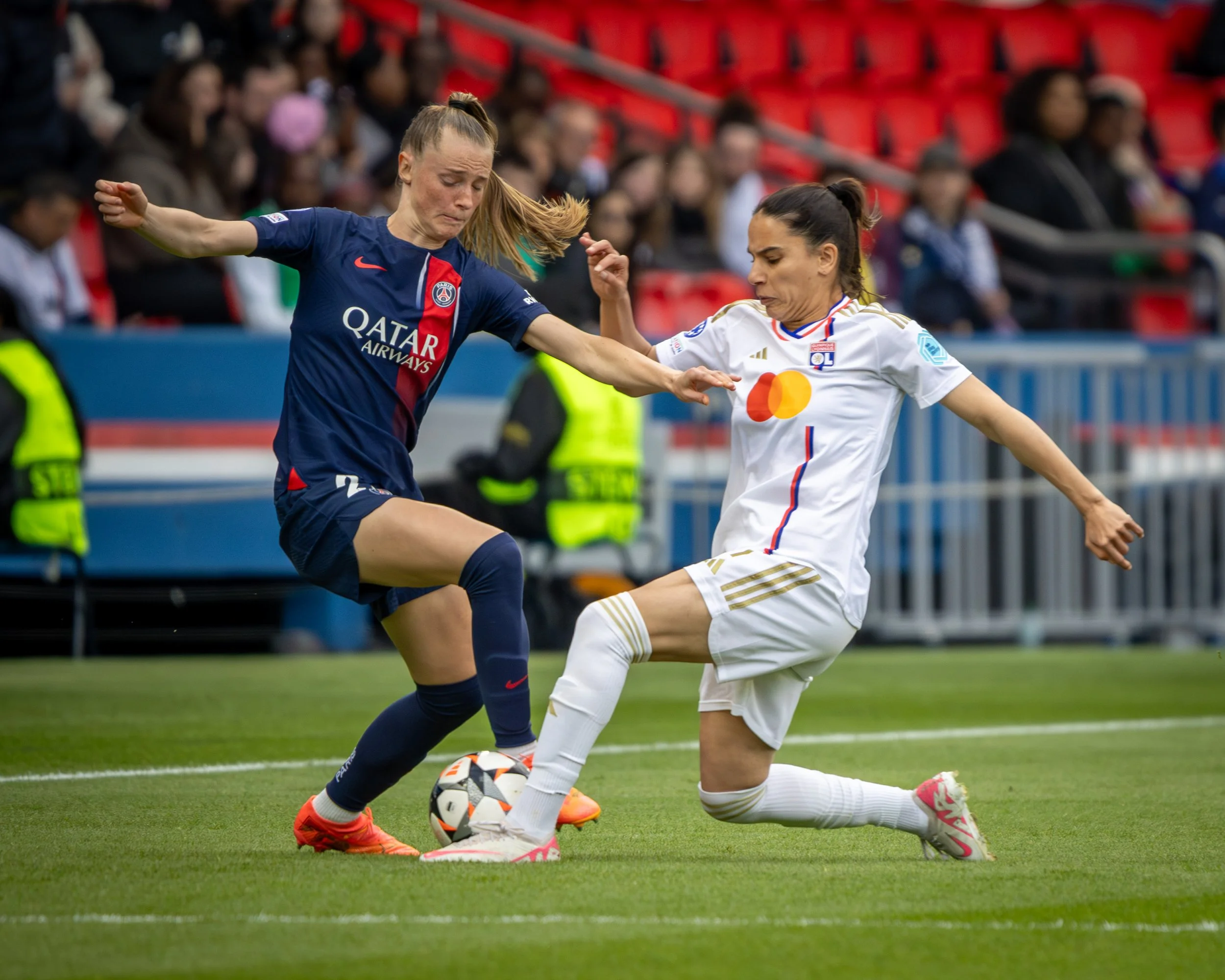 Two women soccer players competing for the ball on the field; one in a blue jersey and the other in a white jersey, with spectators in the background.