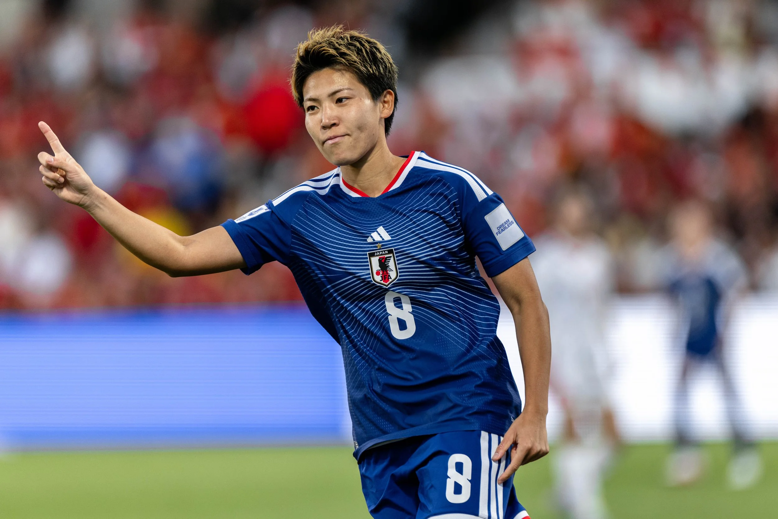 A male soccer player in a blue Japan team jersey celebrating on the field with his right arm raised, pointing upwards with one finger, during a match.