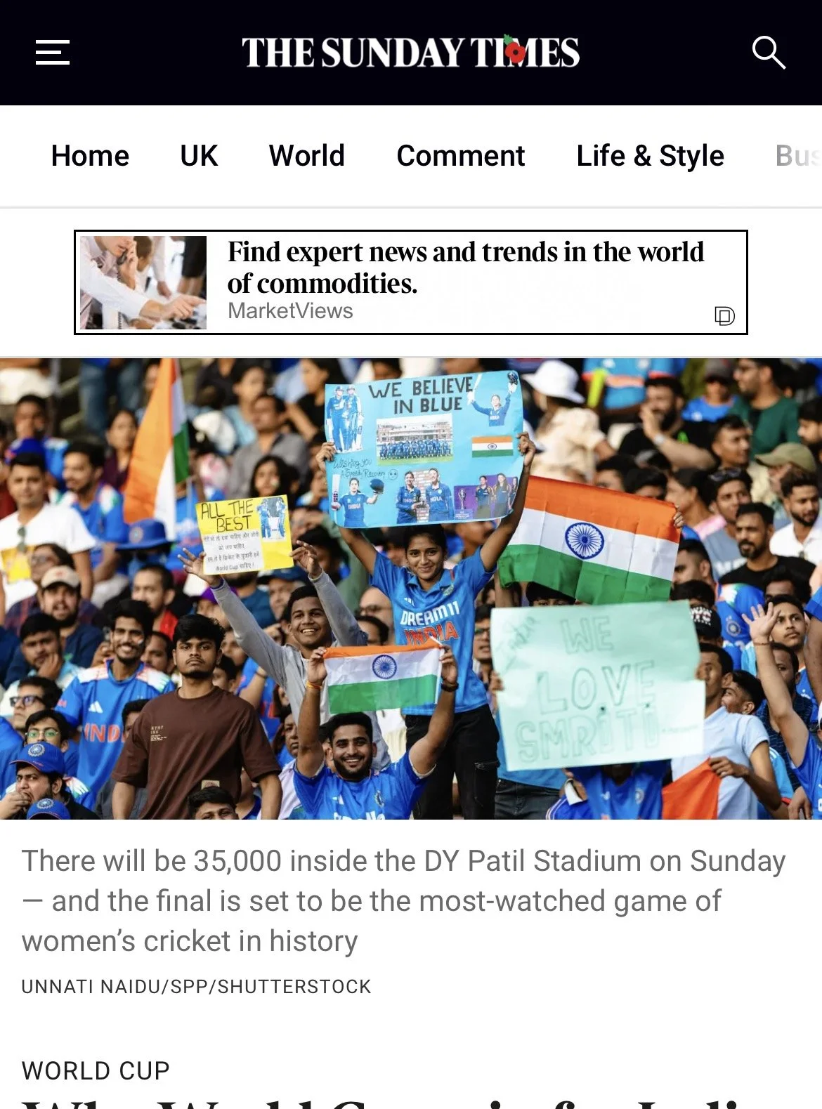 Crowd of fans wearing blue shirts, holding Indian flags and signs in support of the Indian women's cricket team at a stadium.