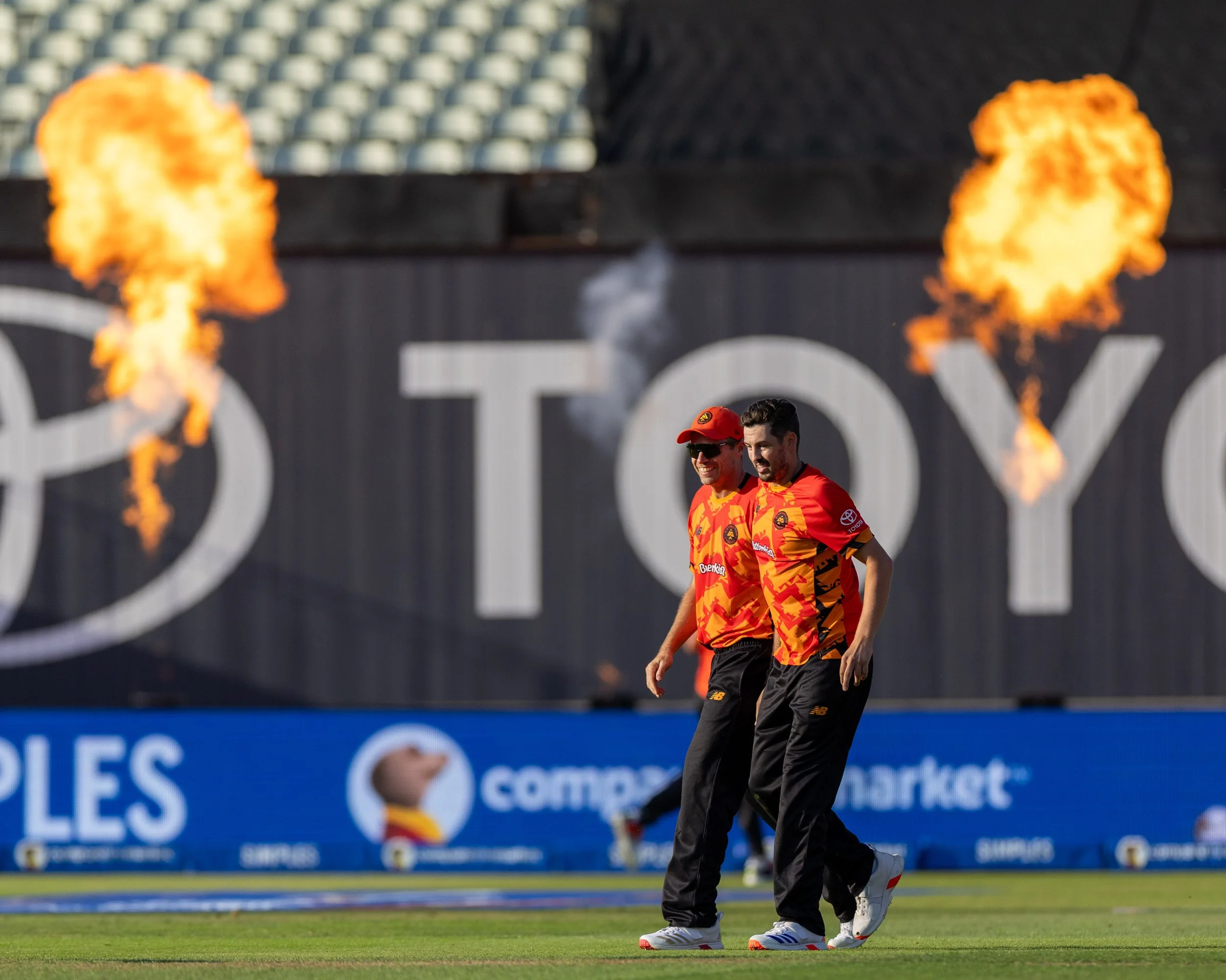 Two soccer players dressed in orange and black uniforms walking on a soccer field with flames over their heads, and a blurred crowd and stadium in the background.