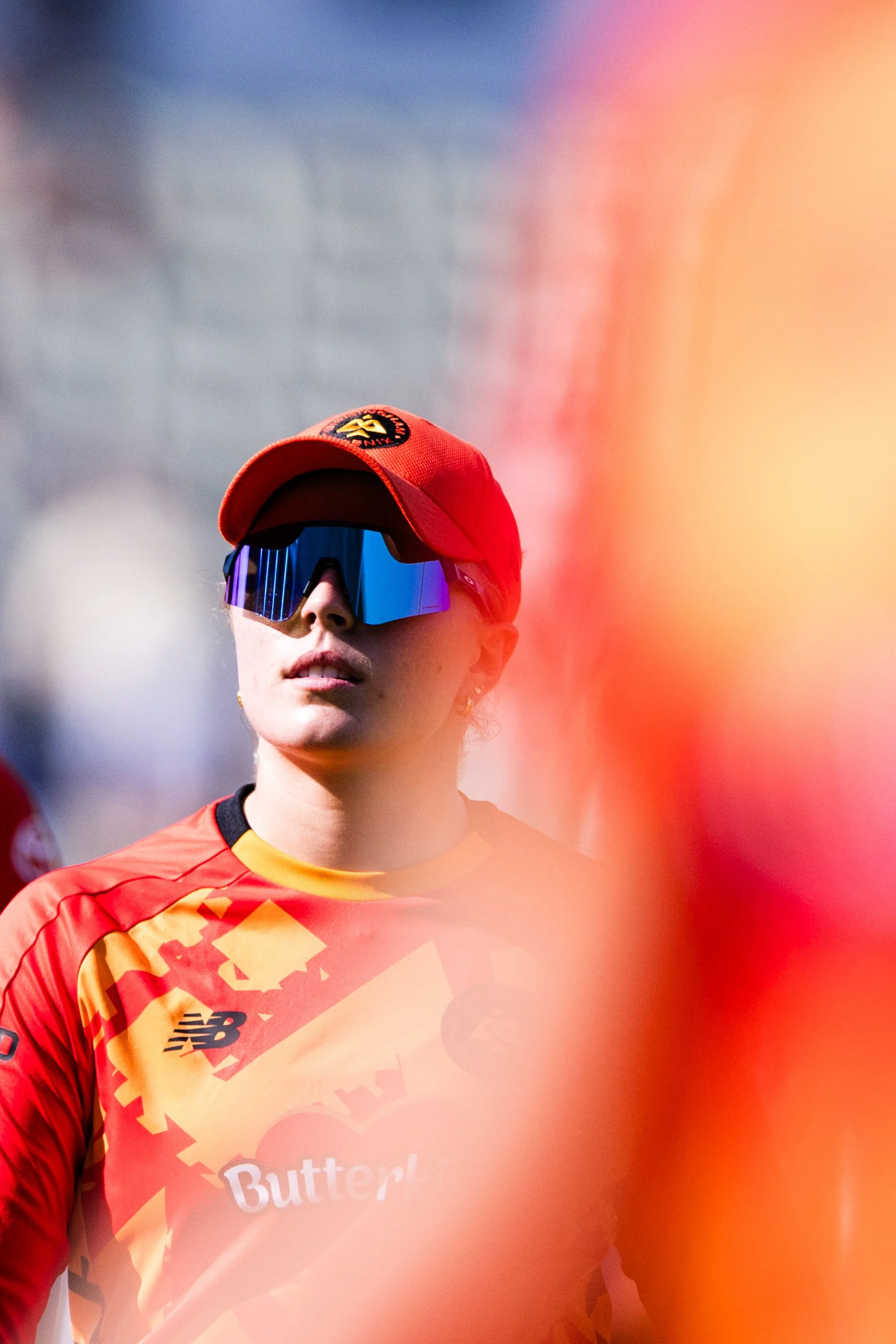 A person wearing a red cap, reflective sports sunglasses, and an orange and red sports jersey with the text 'Butt' and a New York Knicks logo visible, standing outdoors.
