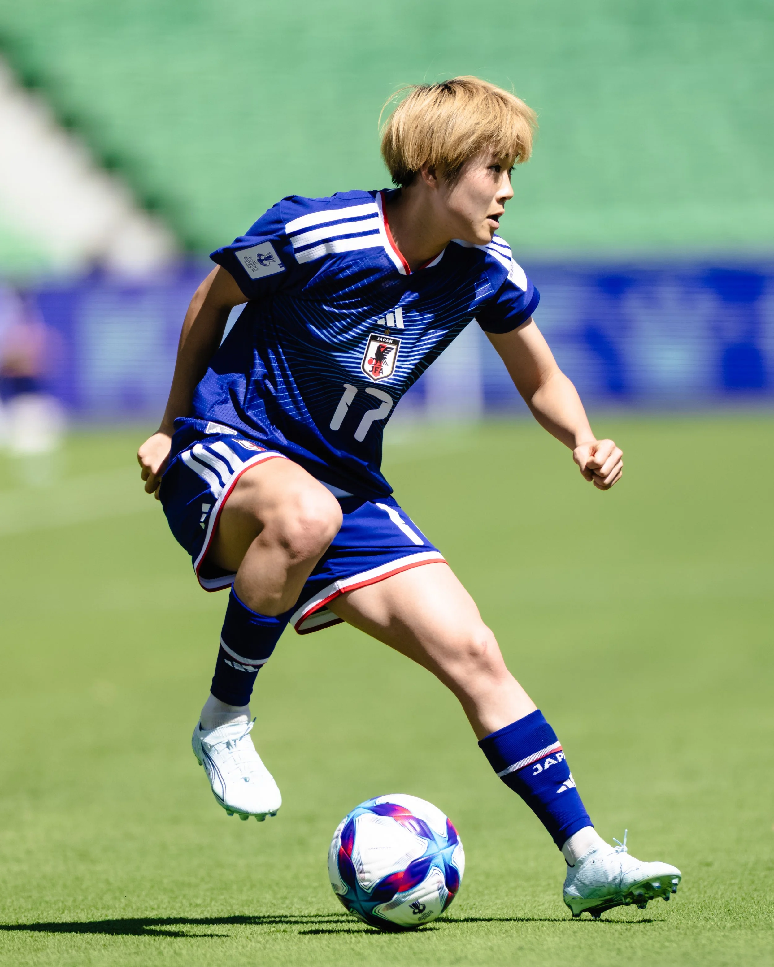 A female soccer player from Japan, wearing a blue jersey with the number 17, is in action on the field, dribbling a colorful soccer ball.