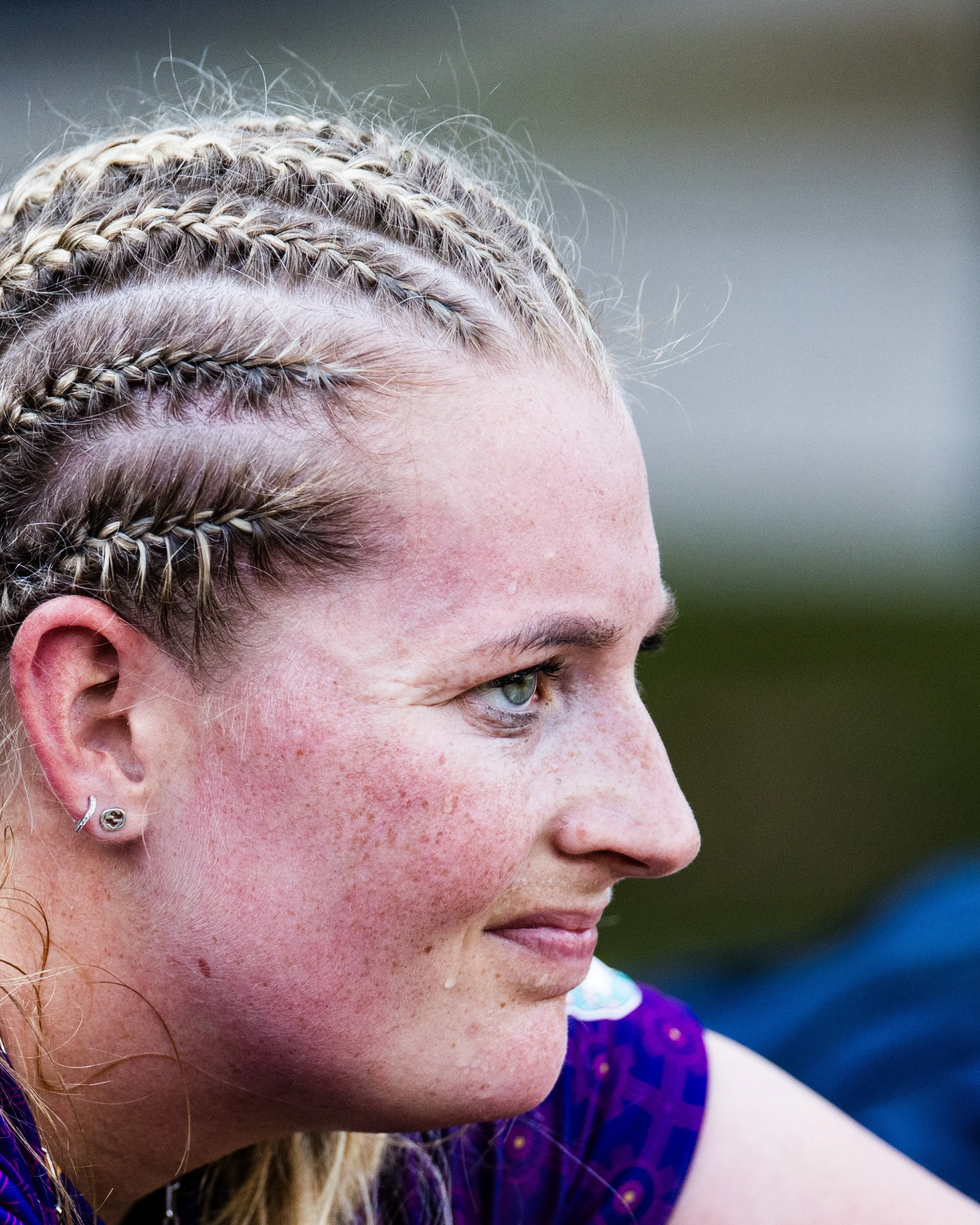 Close-up of a woman's face with blonde braided hair, earrings, and freckles, looking downward, with a blurred background.