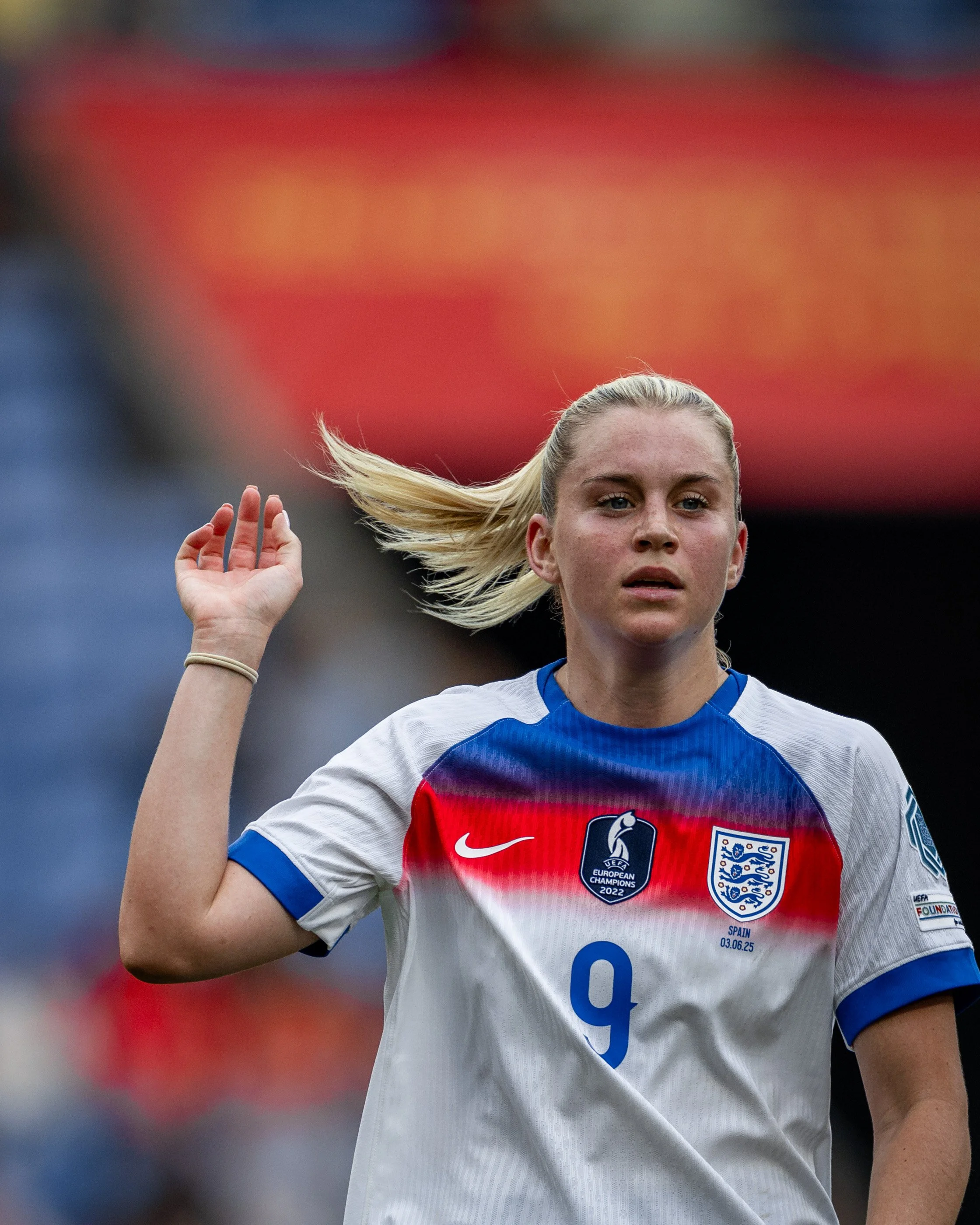 A young female soccer player with blonde hair wearing an England national team jersey raising her hand during a match.