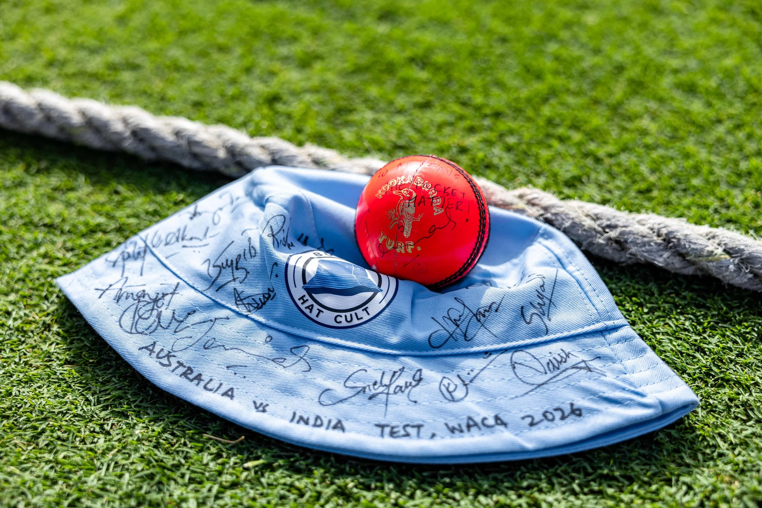 A red cricket ball resting on a white cloth with signatures, placed on a grassy field with a thick rope nearby.