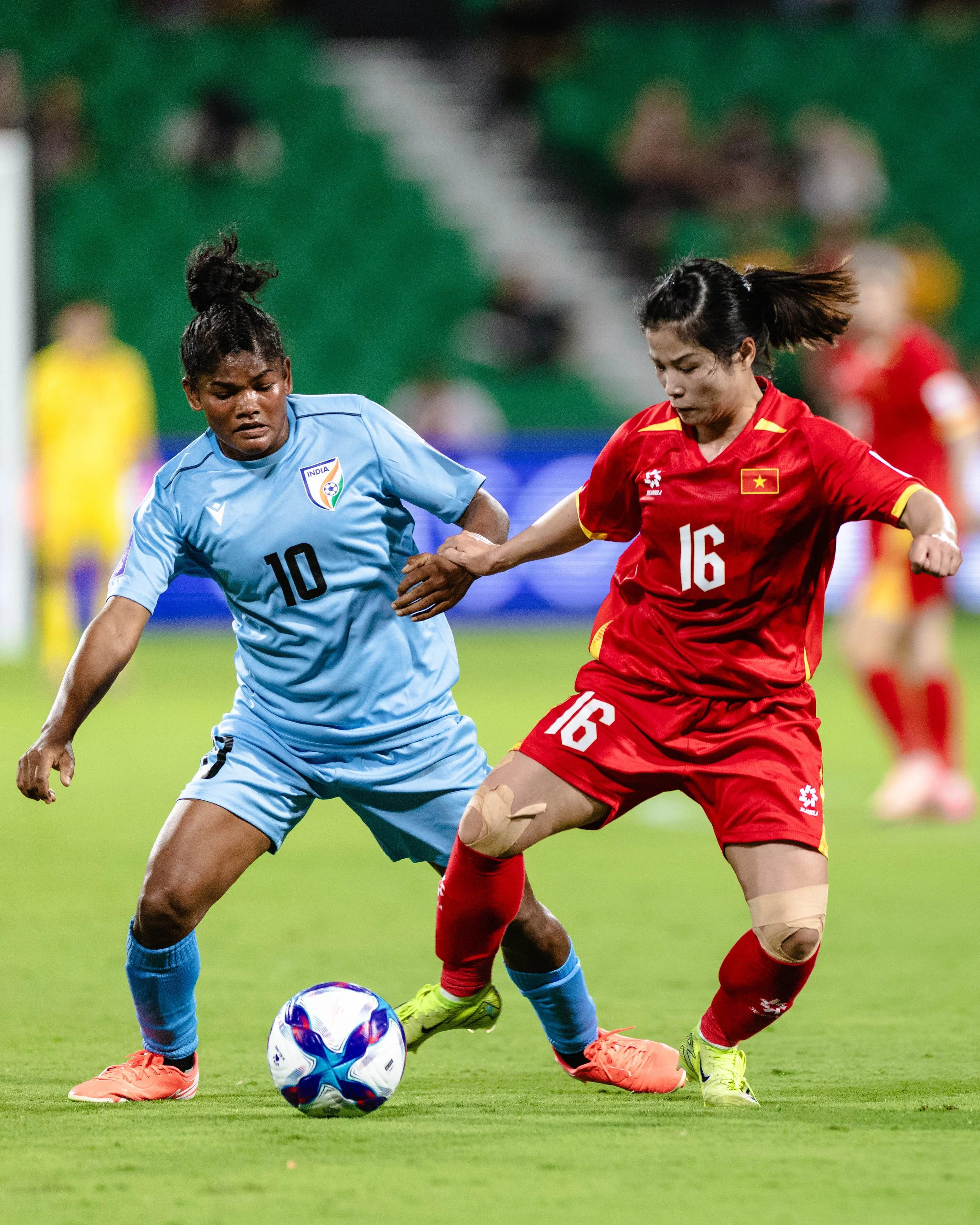 Two female soccer players, one in a blue jersey and the other in a red jersey, compete for the ball on a green field during a match. The player in blue wears number 10 and the player in red wears number 16.