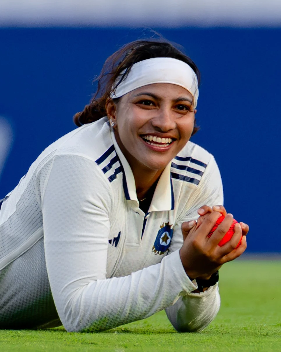 A smiling female cricket player in a white uniform with blue accents, lying on a cricket field, holding a red cricket ball.