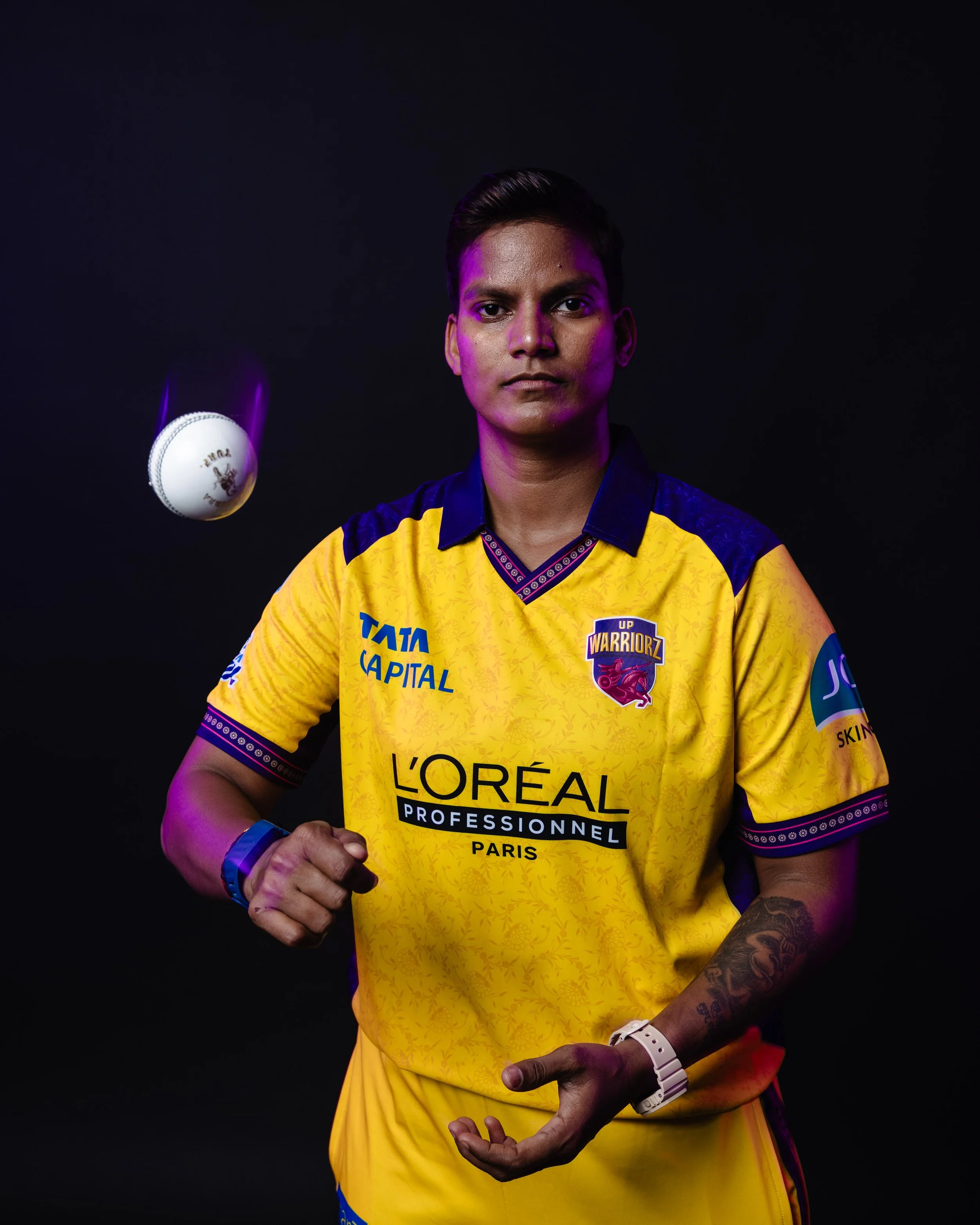 A man wearing a yellow sports jersey with various logos, including 'L'Oreal', 'TATA CAPITAL', and 'UP WARRIORZ', standing against a black background. The man has tattoos on his left arm, is wearing a white watch, and is making a gesture with his hand