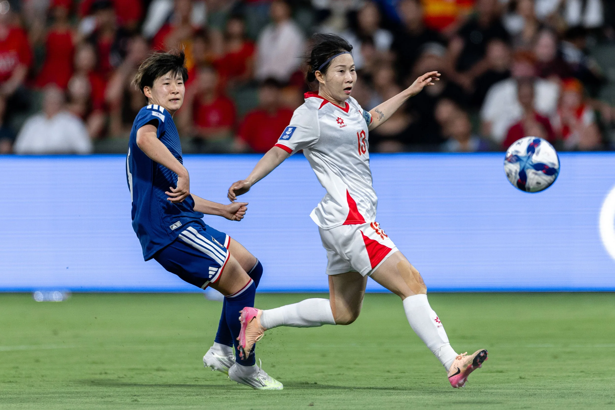 Two female soccer players competing for the ball on the field during a match, with a crowd of spectators in the background.