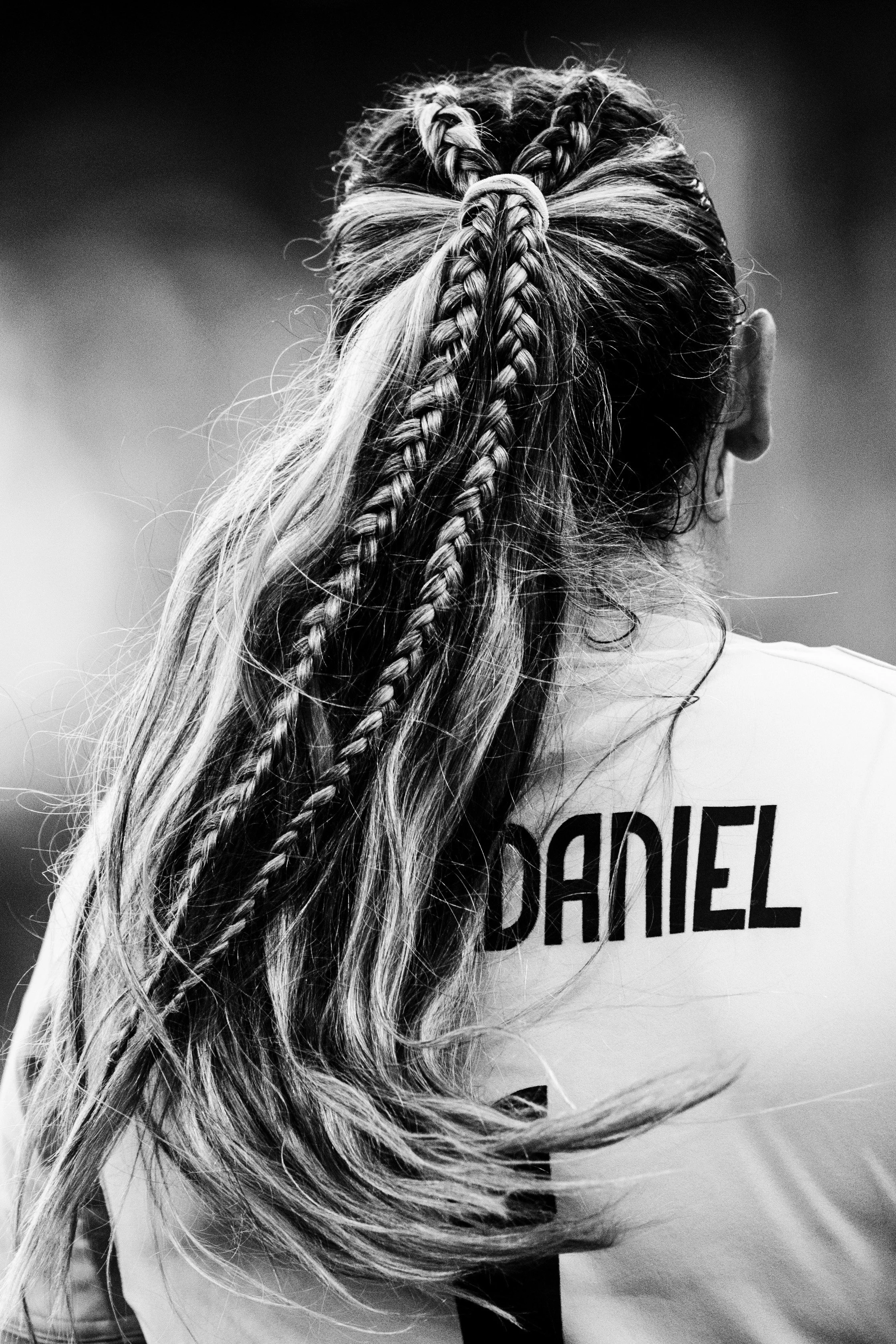 Back view of a woman with long wavy hair styled in multiple braids, wearing a shirt with the word 'DANDEL' partially visible, in black and white.