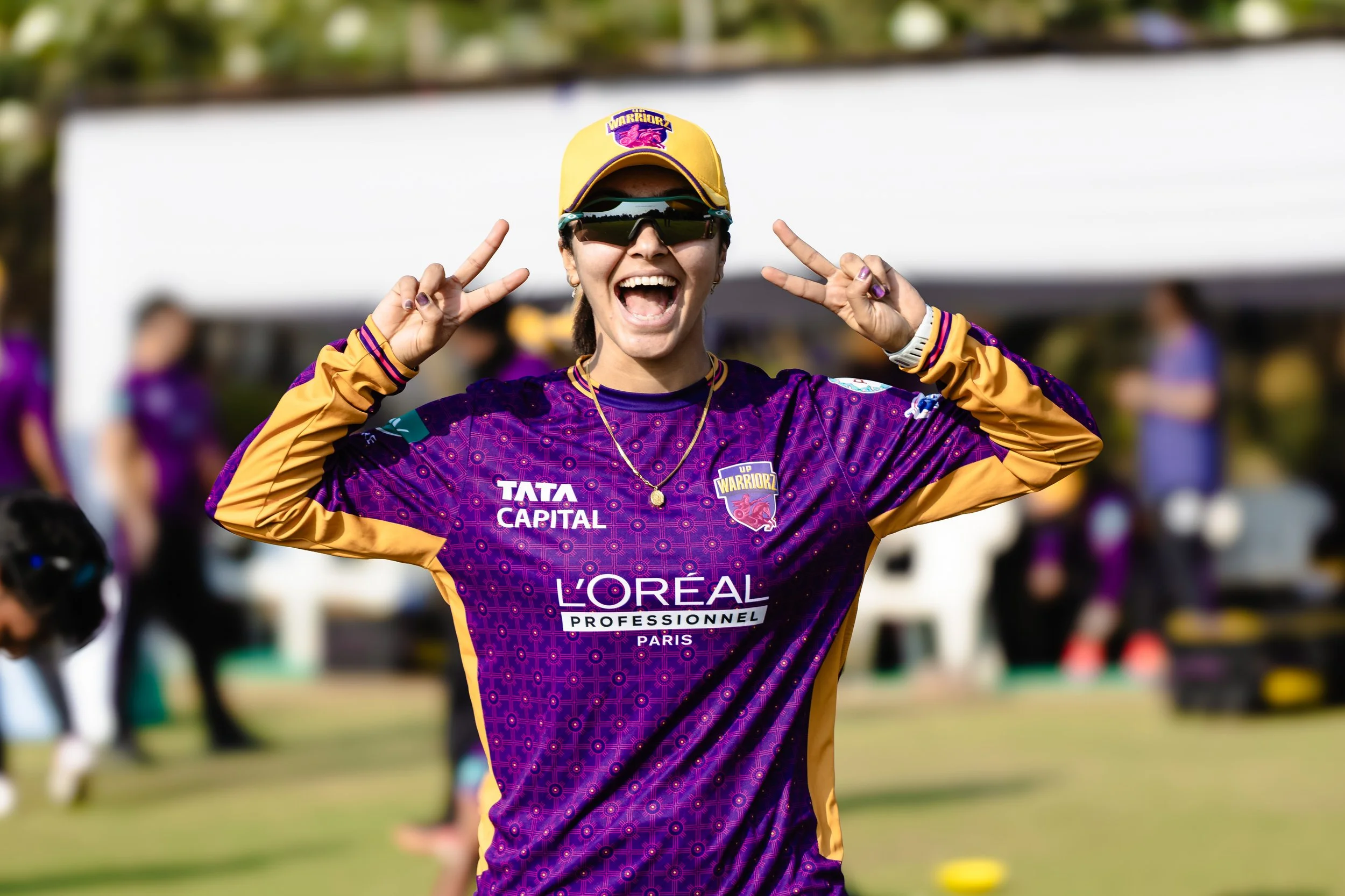 A woman celebrating at a cricket match, wearing sports gear with logos and sunglasses, making peace signs with both hands.