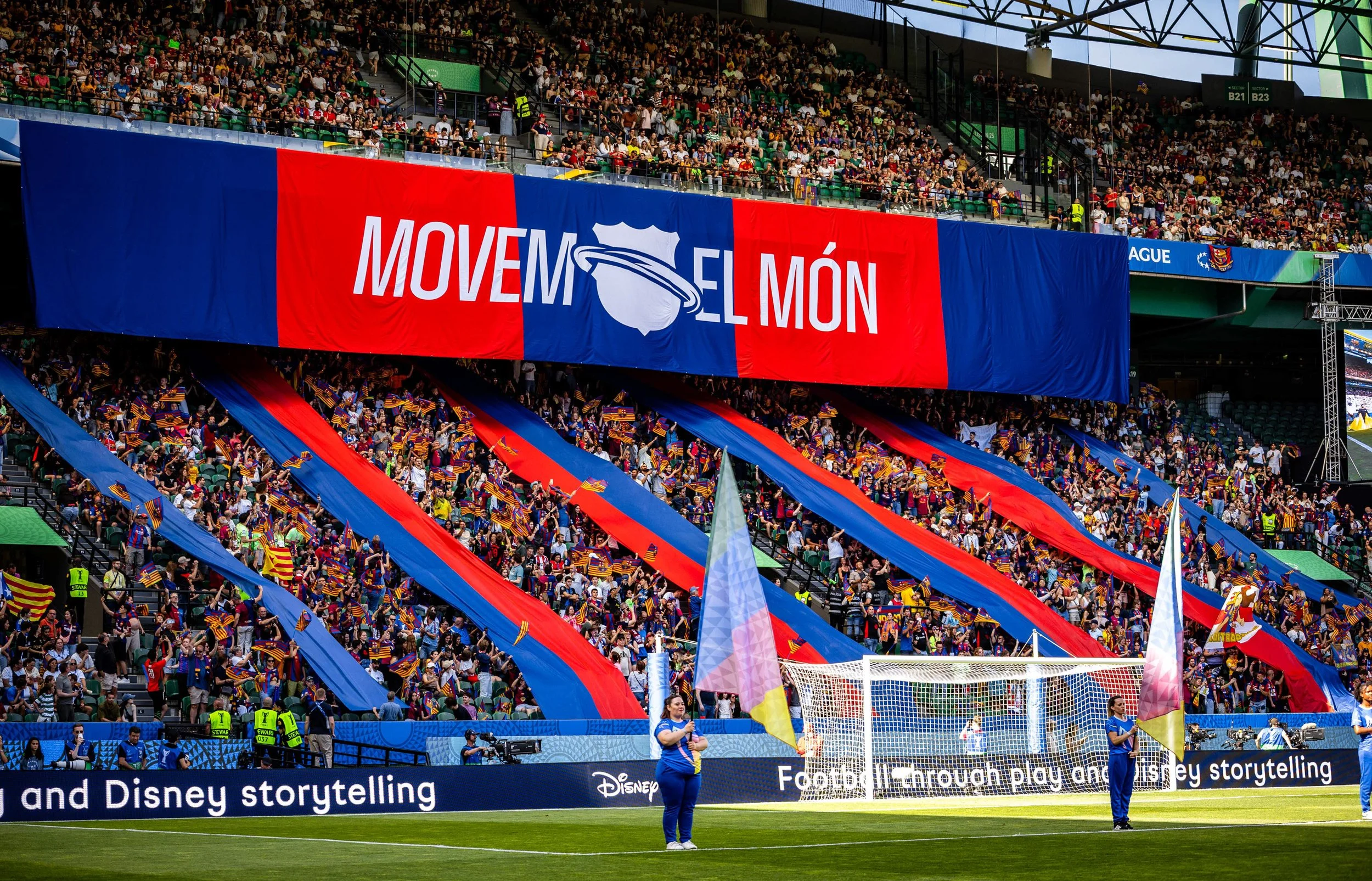 A crowded sports stadium filled with fans, many waving red and blue flags. Large banners display the words 'MOVEM EL MÓN' and a stylized map of the world. The field, with goalposts and a soccer goal, is visible in the foreground, with people standing