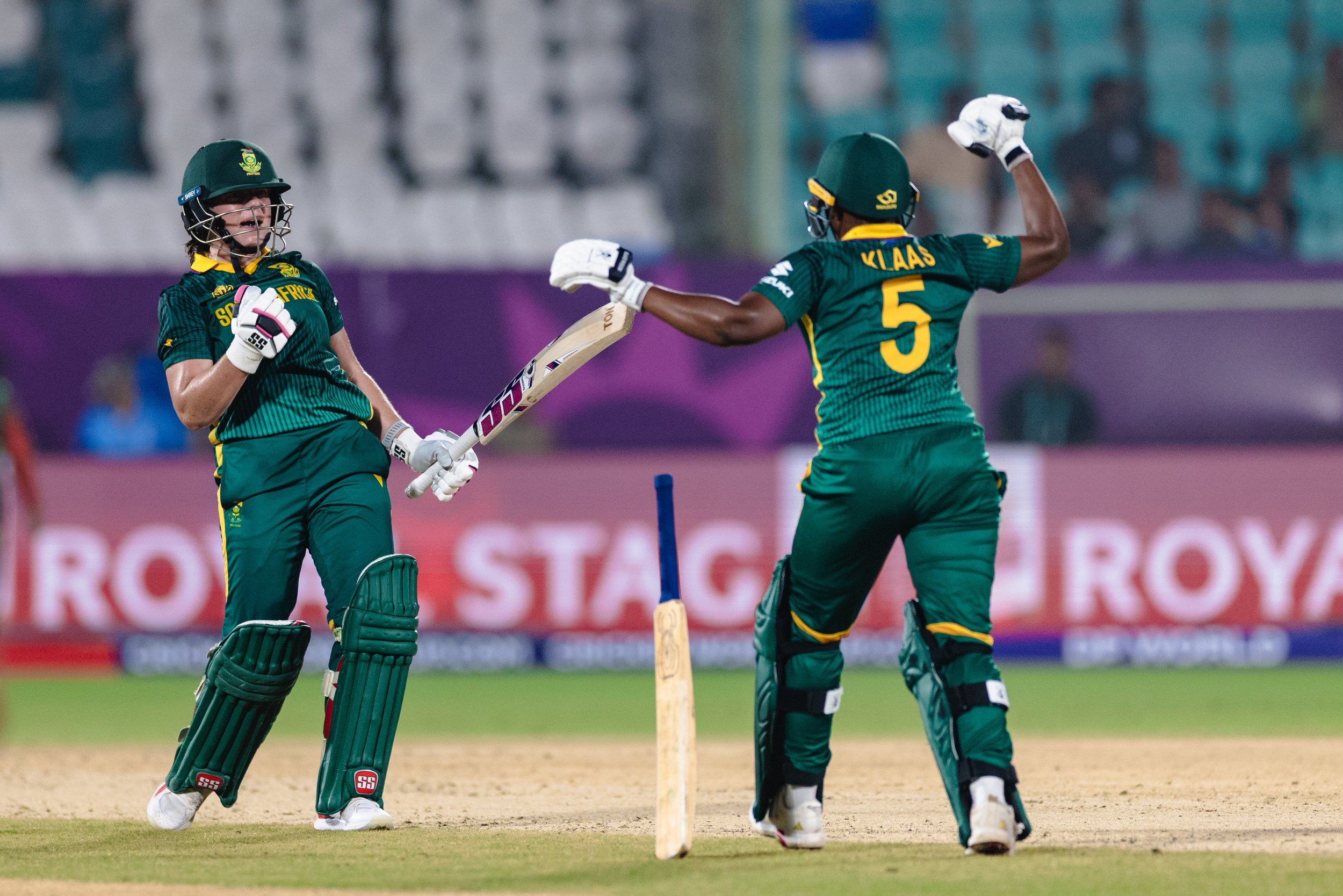 Two cricket players in green uniforms celebrating on the field, with one holding a cricket bat and the other raising a fist.
