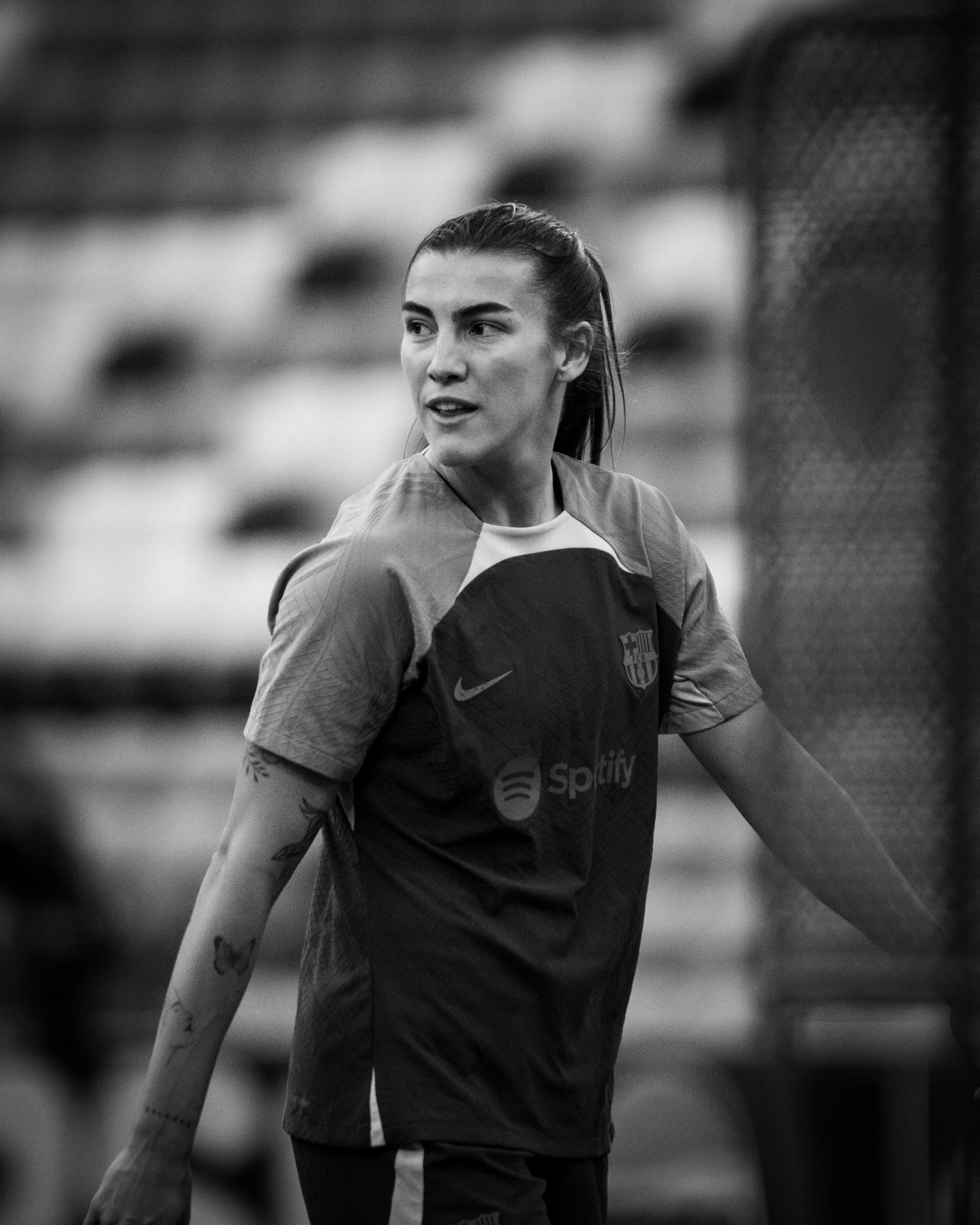 Black and white photo of a female athlete in a sports jersey with a logo, standing outdoors with blurred background, looking to the side.