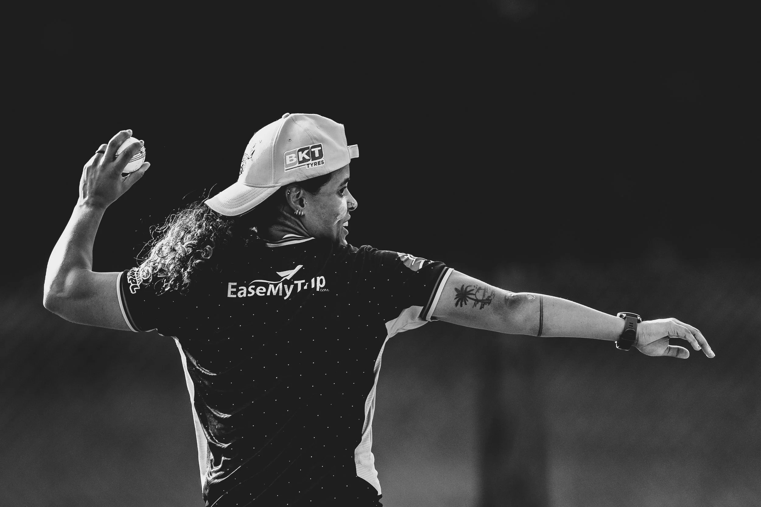A female athlete with curly hair, wearing a cap with the BKT Tires logo, in a black and white photograph. She is holding a ball in her right hand, wearing a sports jersey with sponsorship logos, and extending her left arm outward.