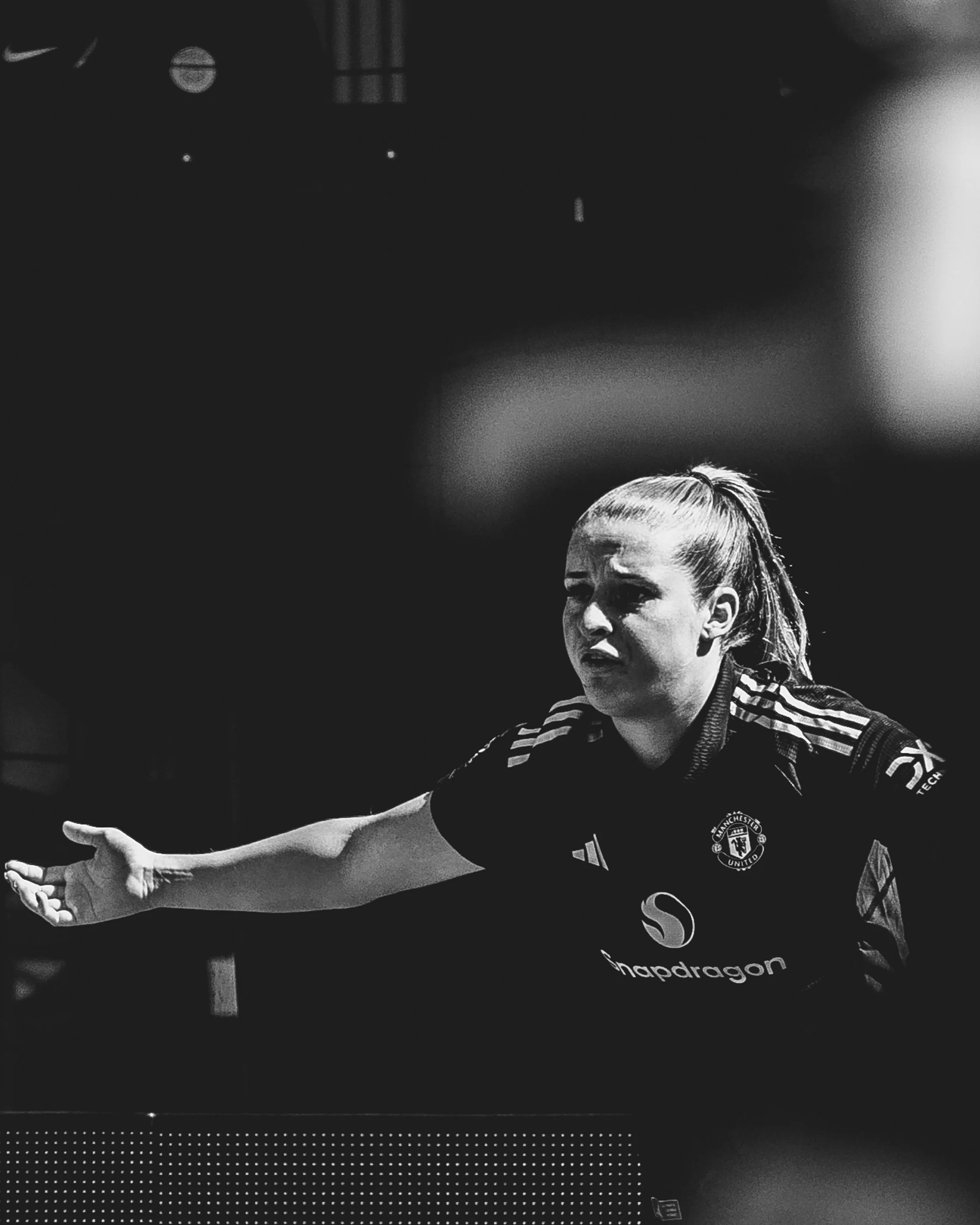 Black and white photo of a female athlete with a bun hairstyle, wearing a Manchester United sports jersey, extending her right arm outward.