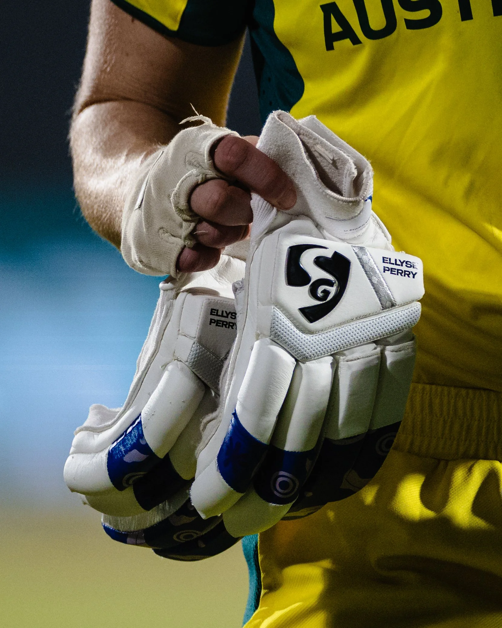 Close-up of an Australian cricket player, wearing a yellow jersey, white wicketkeeping gloves, and holding a white cricket ball, with a blue background.