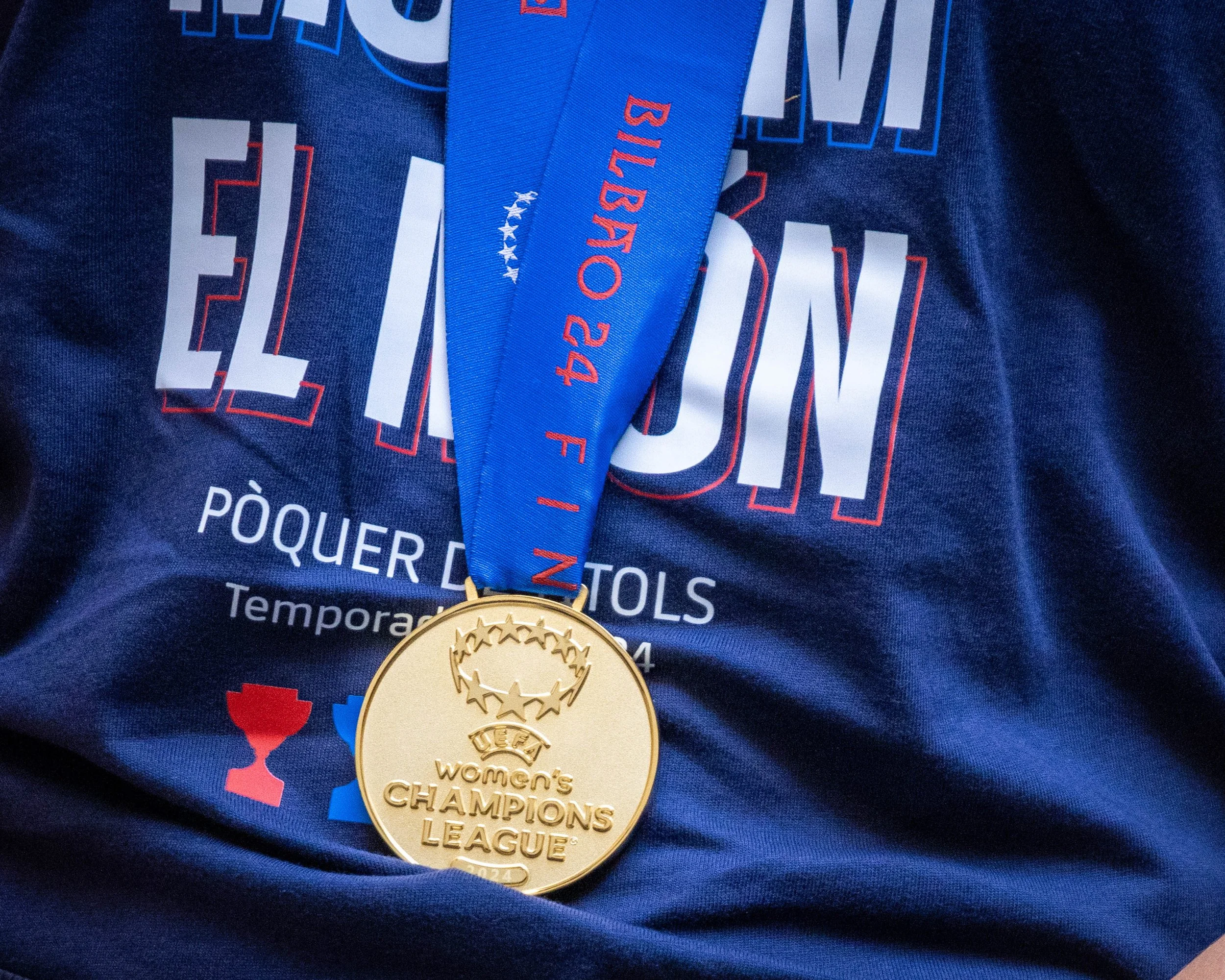 Close-up of a gold medal with the text "Women's Champions League" hanging around a person's neck, with a blue ribbon. Part of a dark blue t-shirt with white and red printed text and graphics is visible.