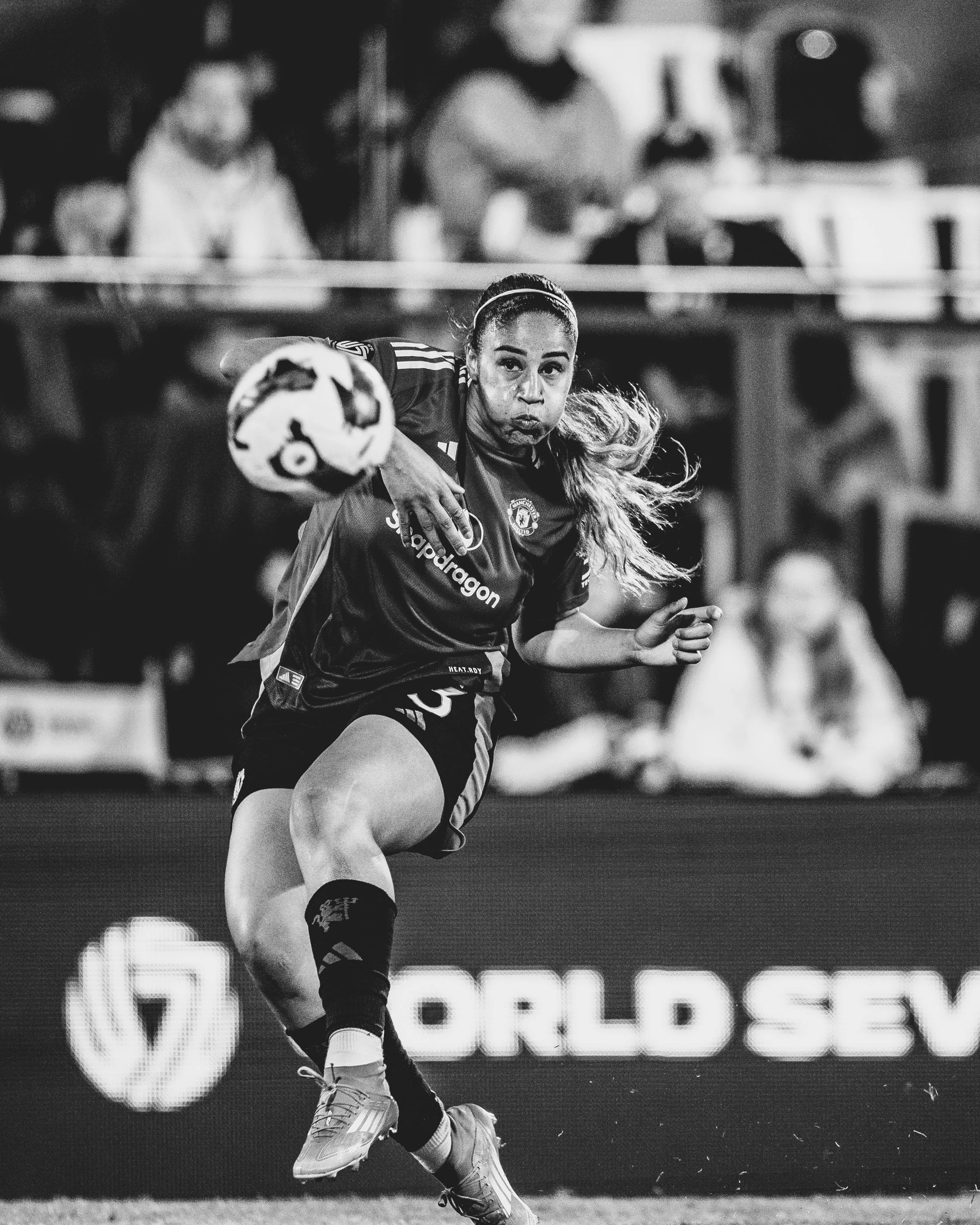 A female soccer player in action during a game, wearing a jersey with sponsorship logos, black socks, and gray cleats, holding a soccer ball while running. The background is blurred with spectators and advertisements.