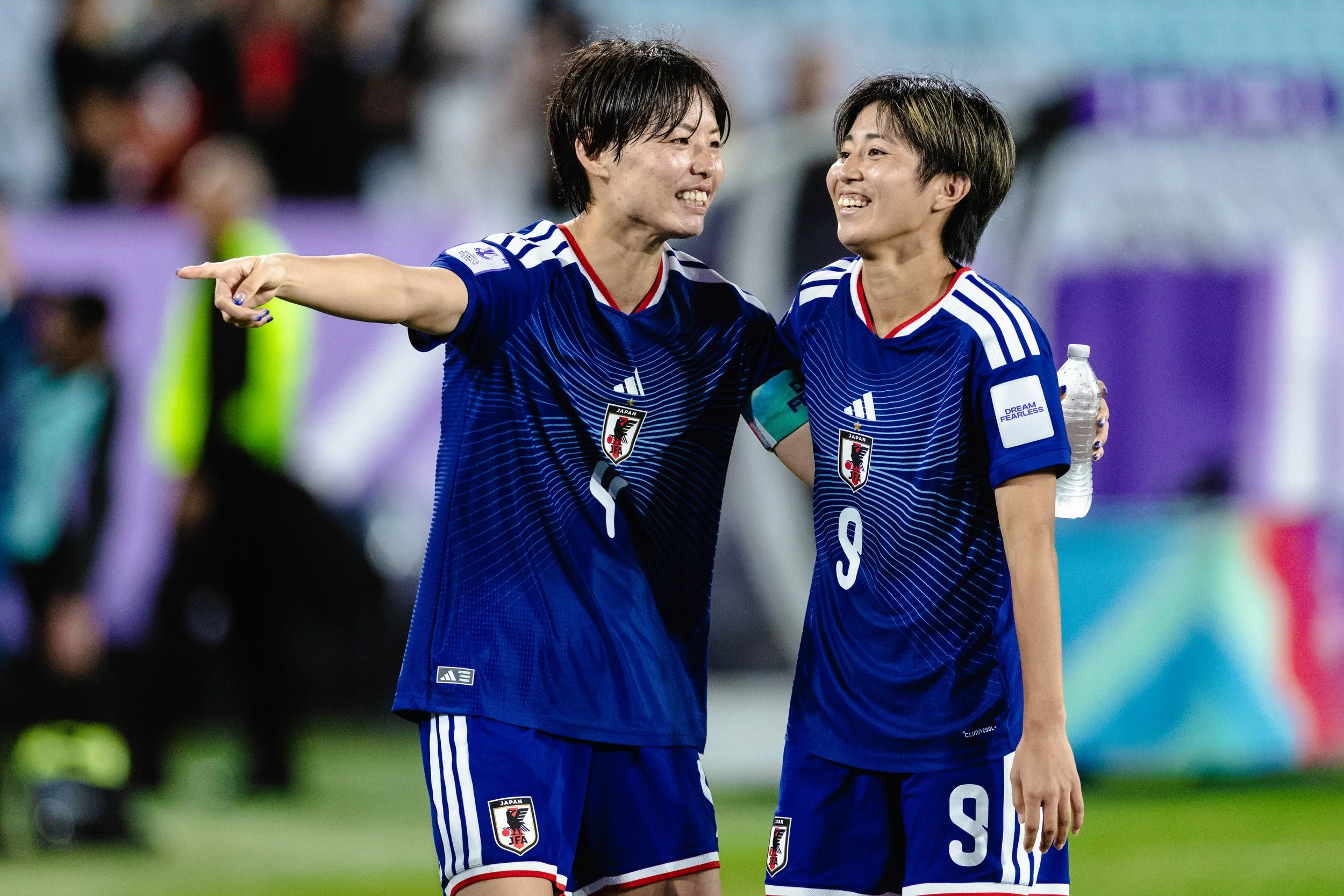 Two female soccer players in Japan national team uniforms smiling and celebrating on the field, one with arm around the other, holding a water bottle.