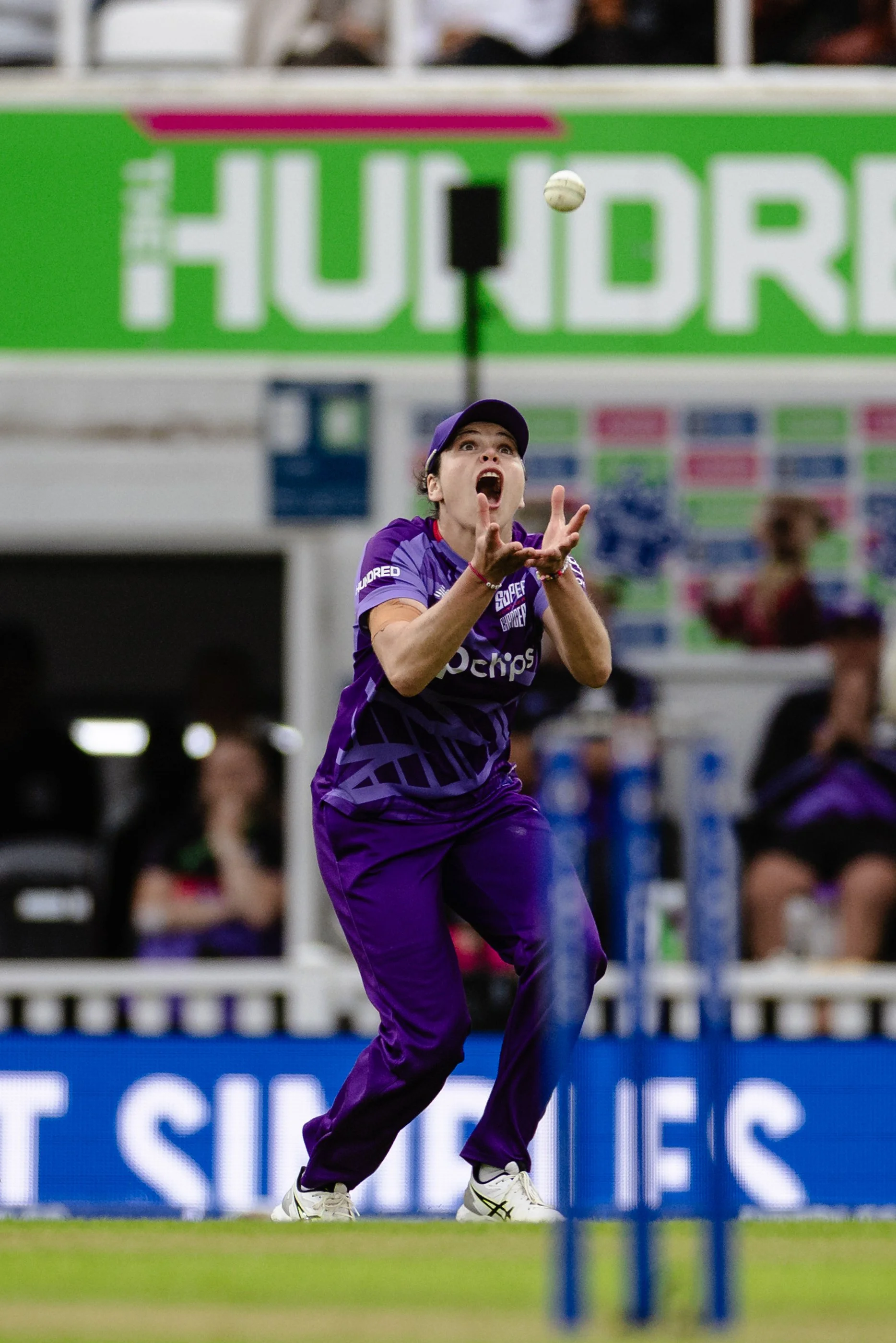 A female tennis player in a purple uniform and cap hitting a tennis ball, with a green sign and spectators in the background.