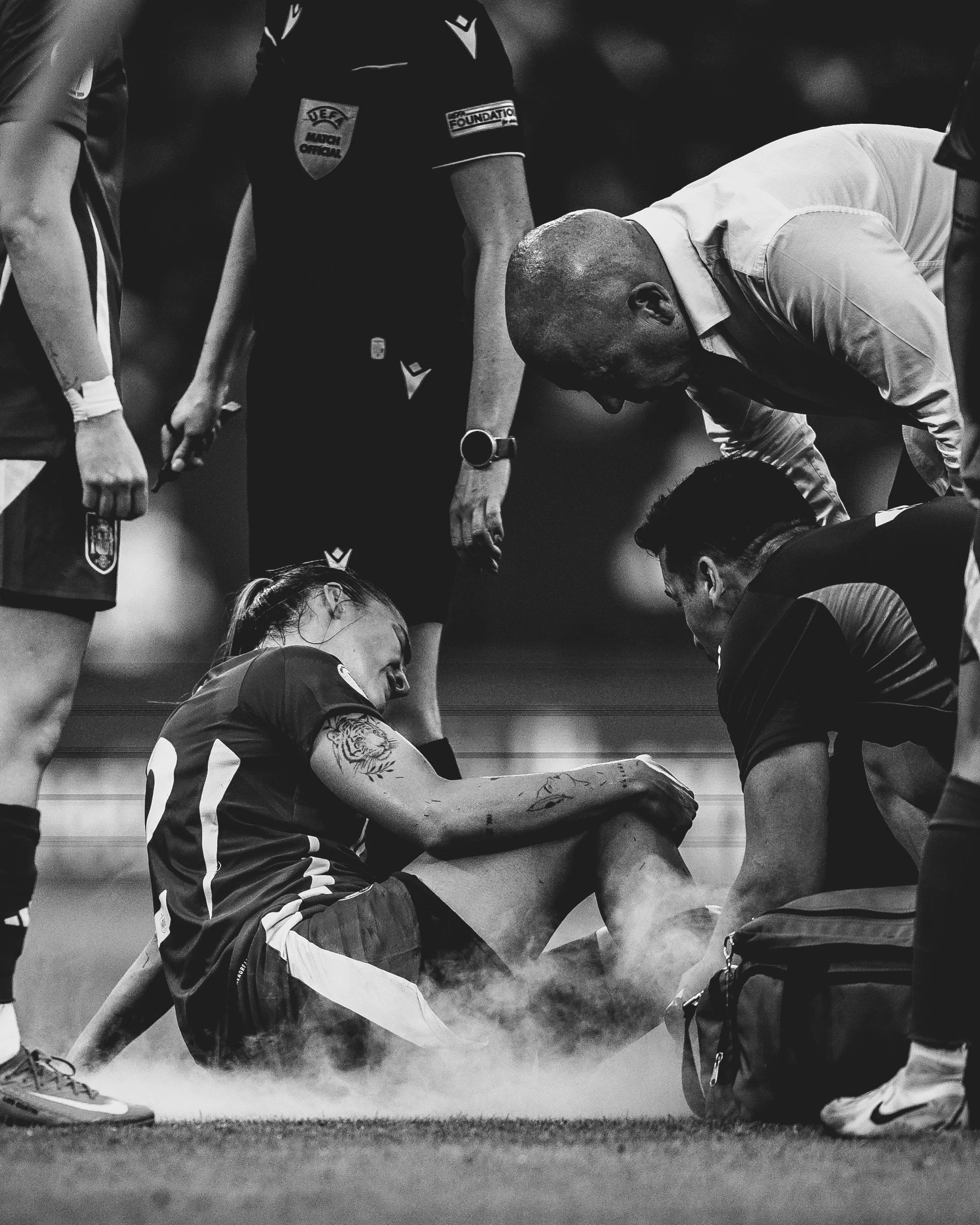 A black and white photo of a female athlete receiving medical attention on the sideline during a sports game, with others standing nearby.