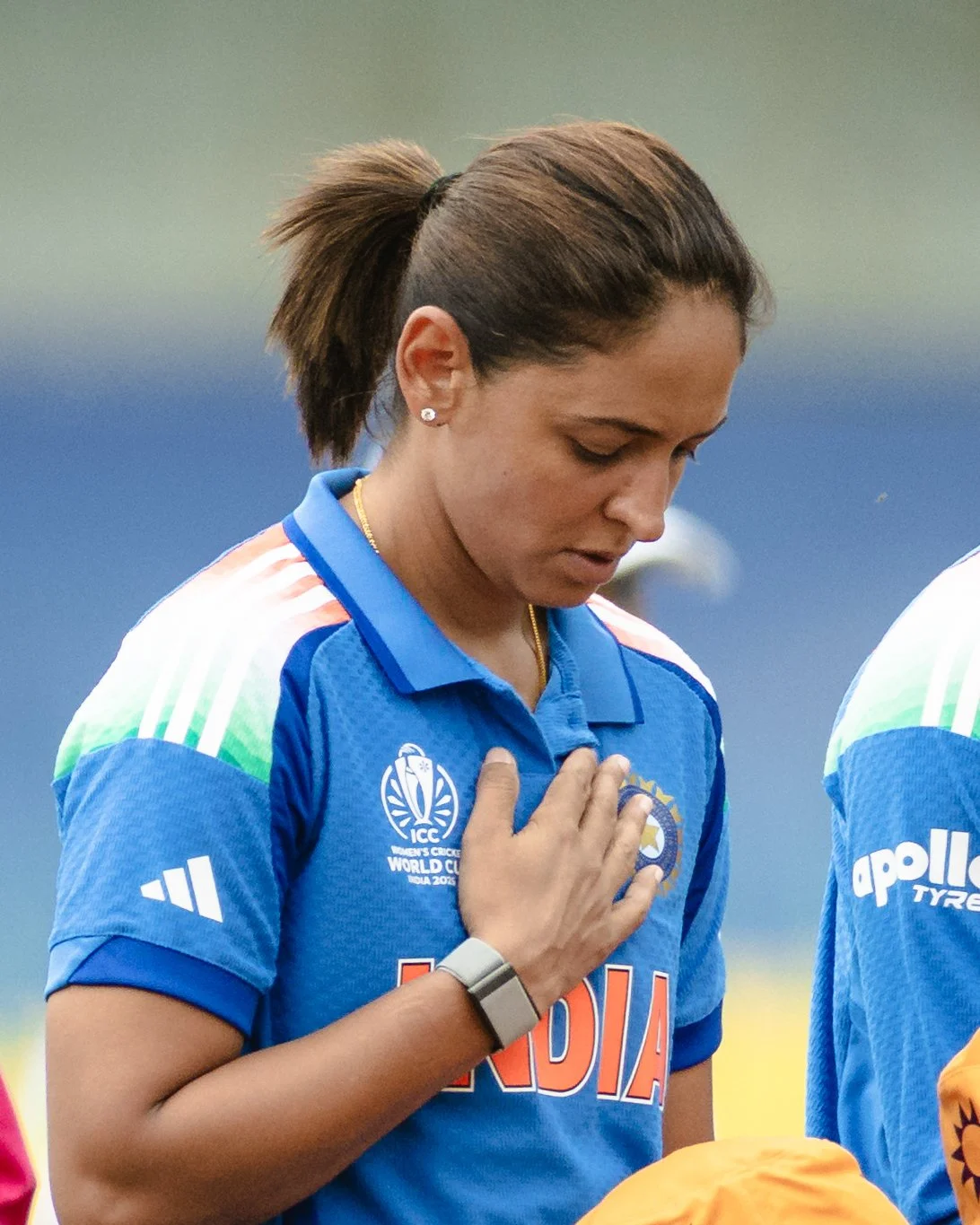 A female cricket player in a blue India team uniform with her hand on her heart, head bowed, during the national anthem.
