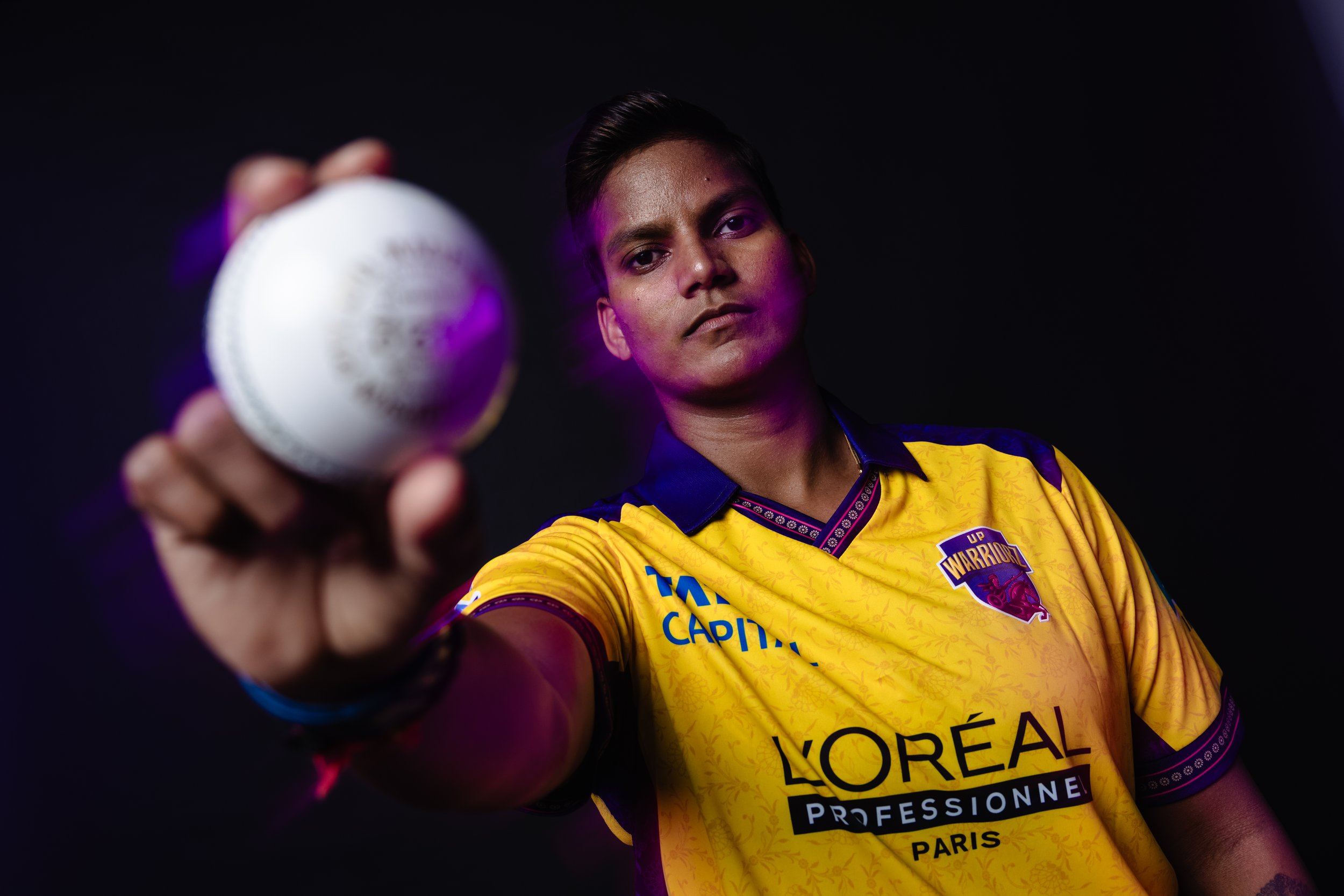 A rugby player wearing a yellow and purple jersey, holding a rugby ball towards the camera, with a black background.