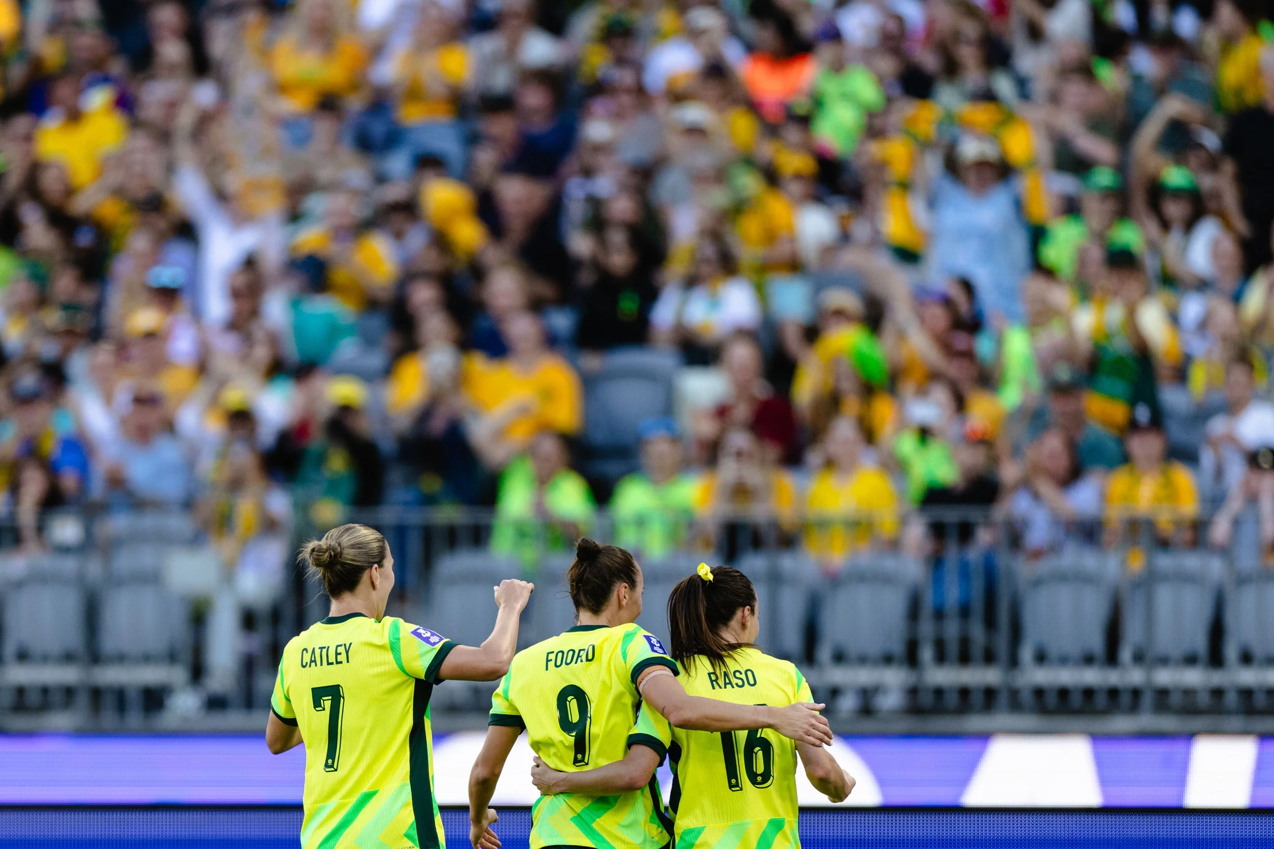 Women soccer players in yellow jerseys with green accents walking on the field during a game, with a blurred crowd of spectators in the background.