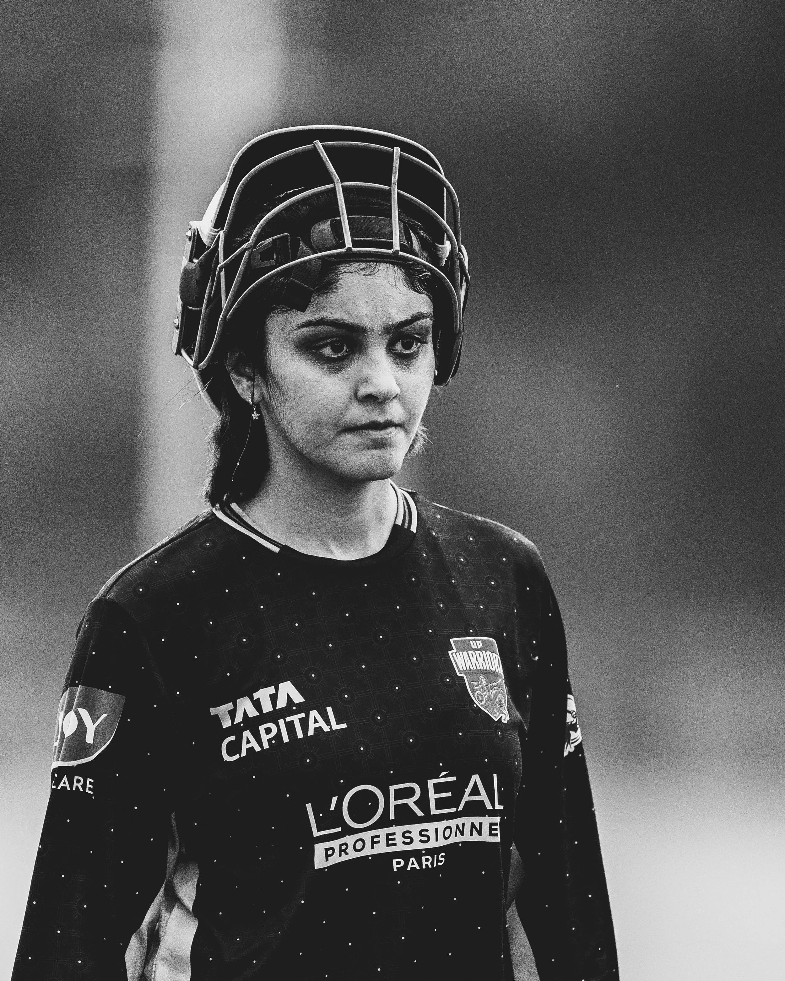 Close-up of a female athlete wearing a sports helmet and jersey with sponsor logos in black and white. She has dark hair, a focused expression, and is captured in a moment of concentration.
